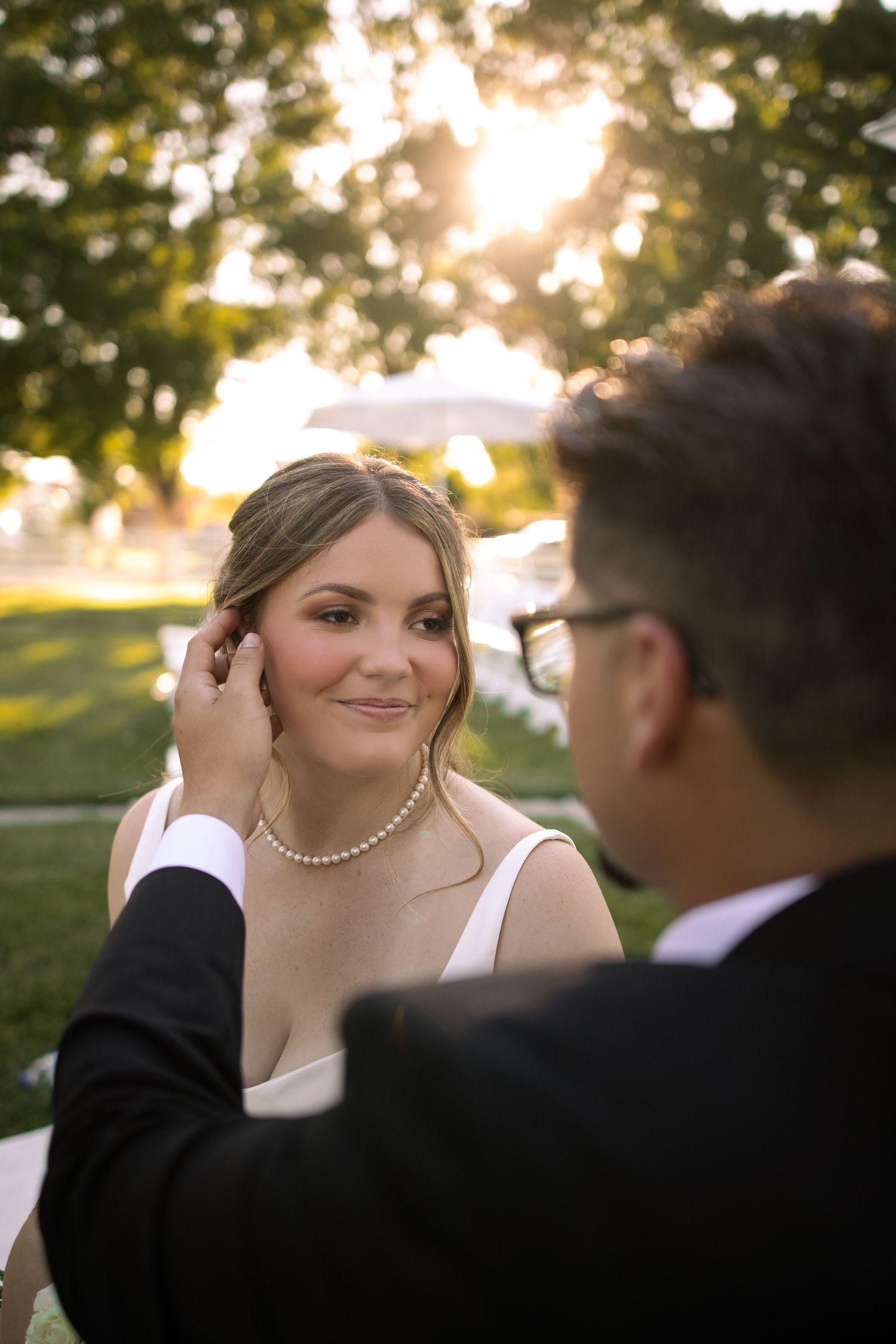 Groom tenderly adjusts brides pearl earring outdoors at golden hour, bride smiling softly in white dress.