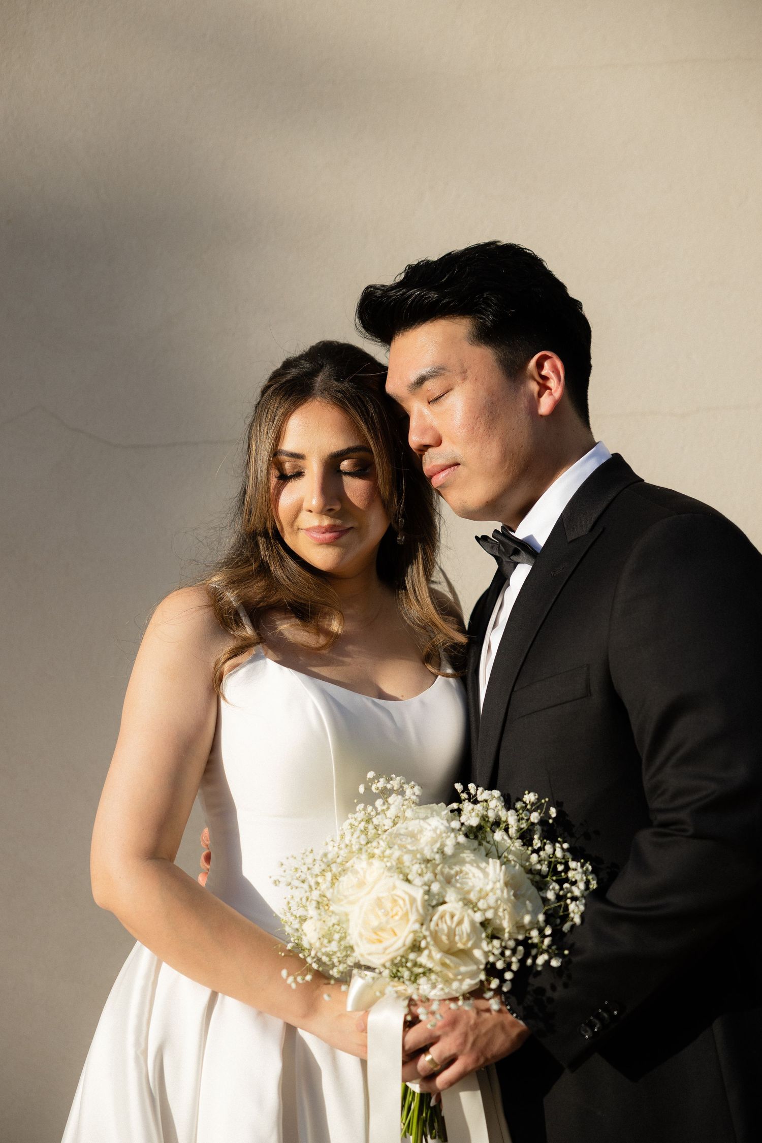 Elegant wedding couple posing together, bride in white gown holding white roses bouquet, groom in black tuxedo with bow tie.