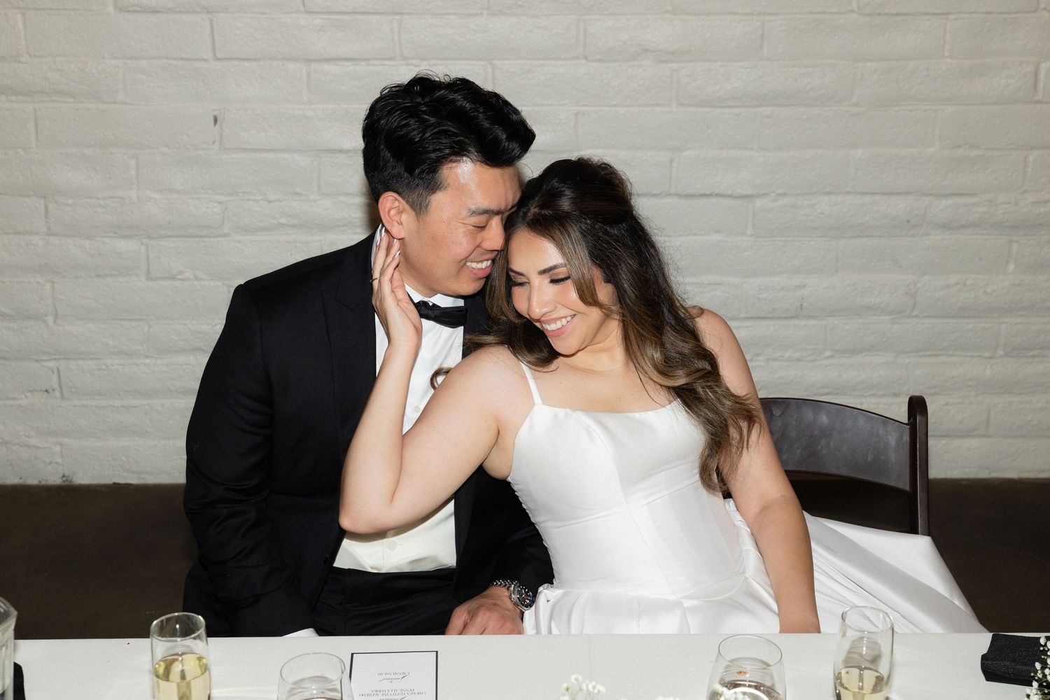 Newlyweds share intimate romantic moment seated at wedding reception sweetheart table with champagne glasses and white brick backdrop.