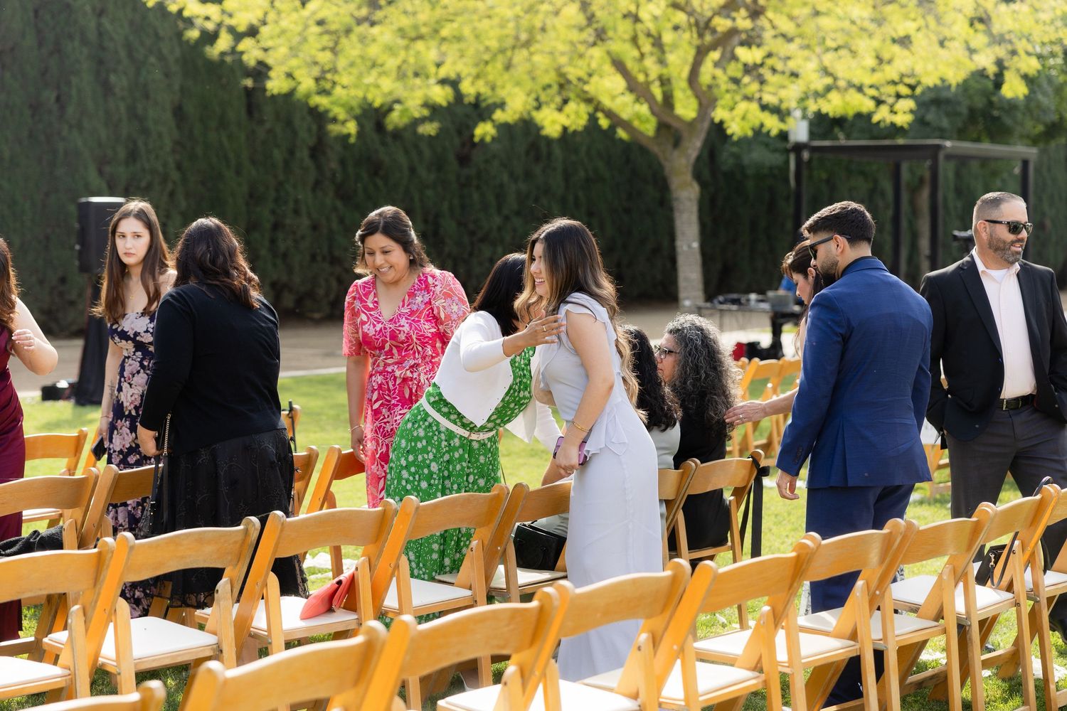 Guests mingle at an outdoor wedding ceremony with wooden chairs on a sunny day.