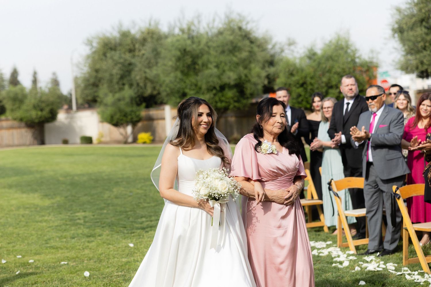 Bride in white gown walks down outdoor aisle with woman in pink dress at sunny wedding ceremony.