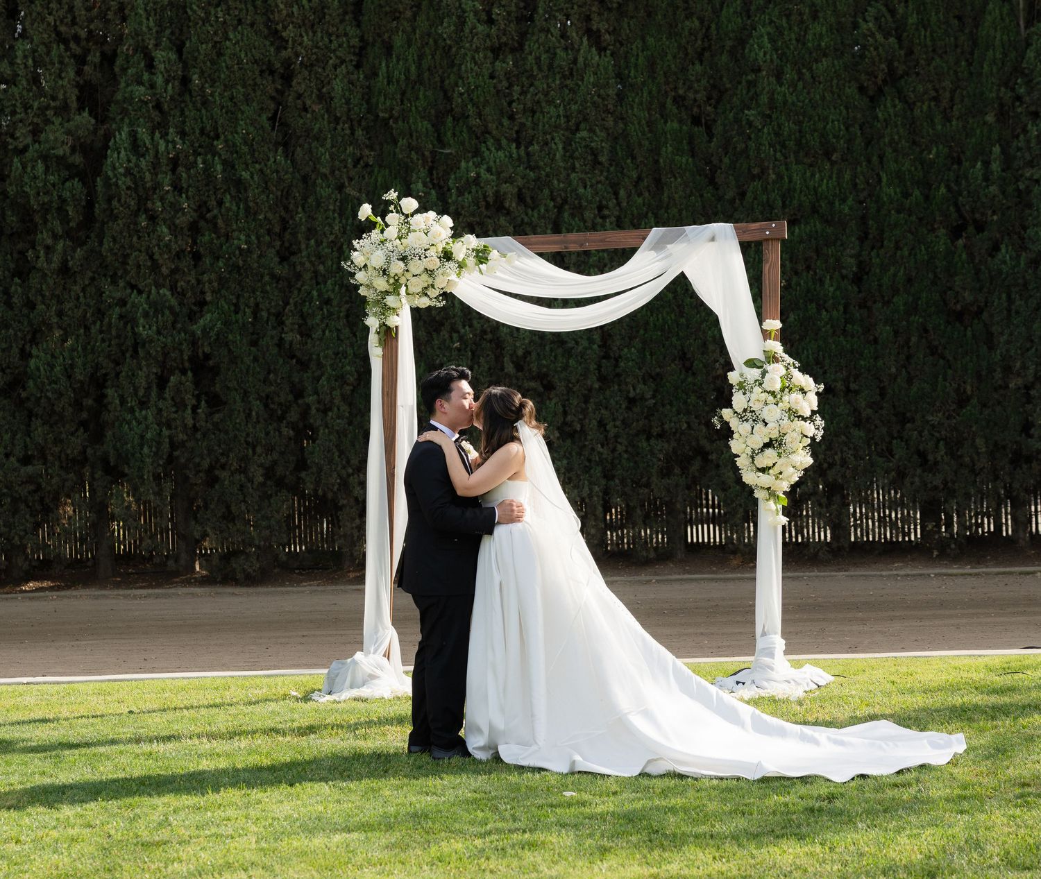 Bride and groom share first kiss under white draped floral arch at outdoor garden wedding.