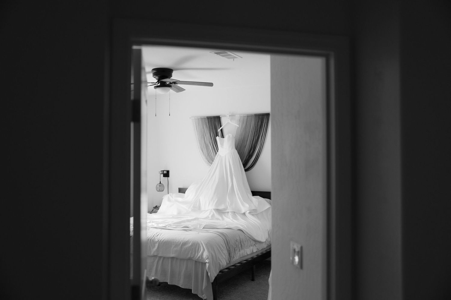 Black and white photo of a wedding dress hanging in a sunlit bedroom, viewed through a doorway.