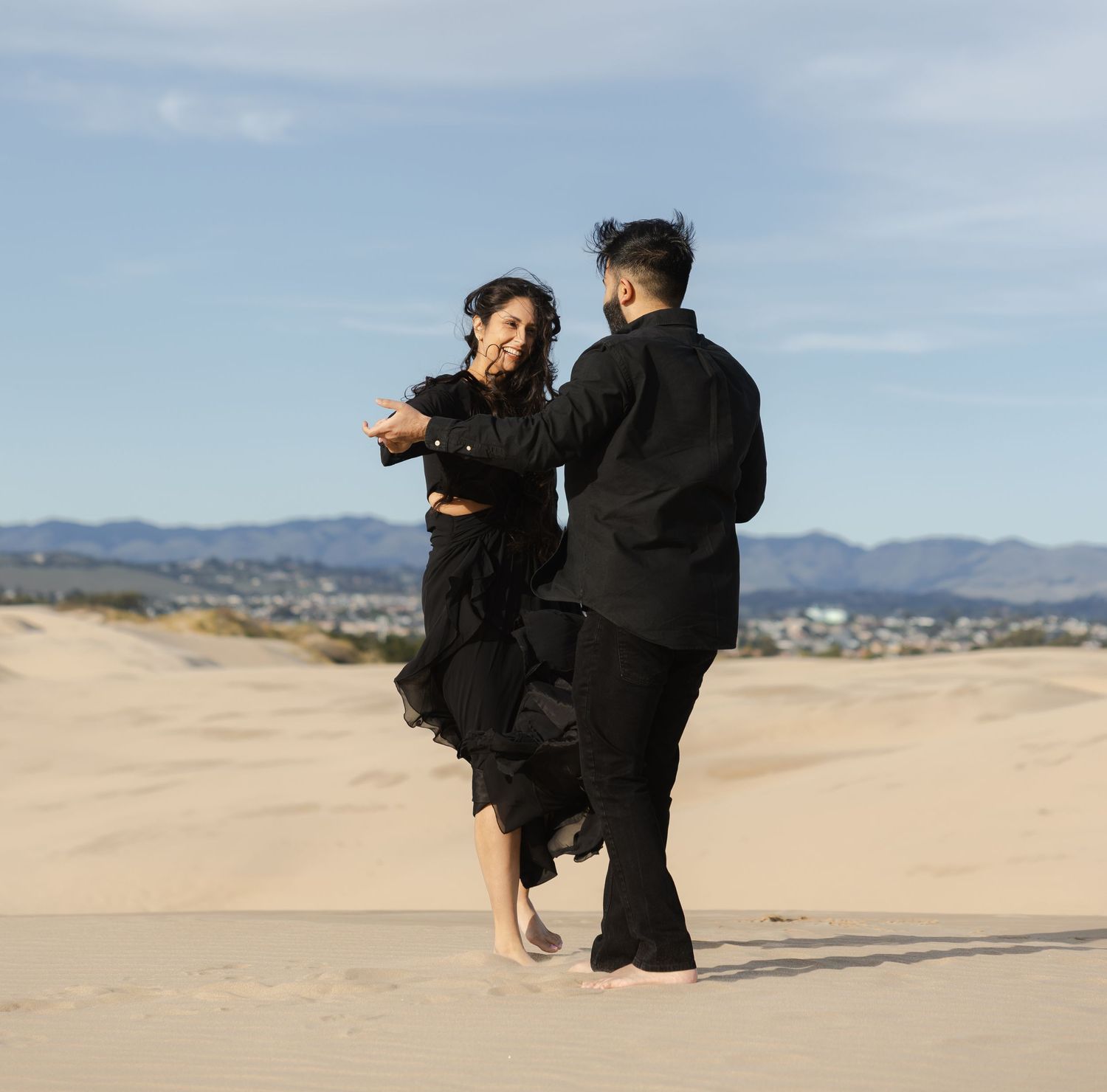 Person in black martial arts attire performs a high spinning kick in a desert landscape with mountains in the distance.
