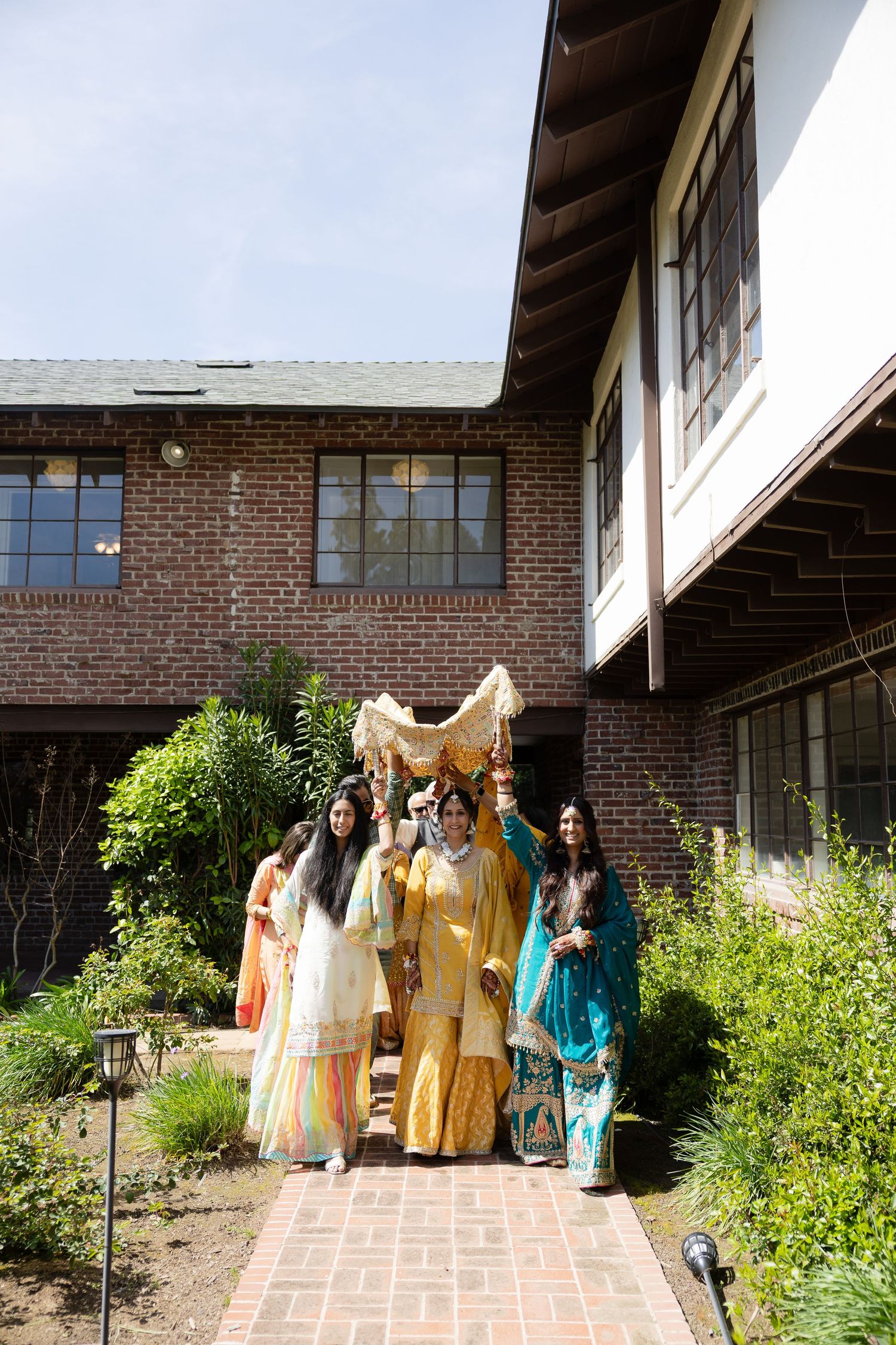 Wedding party members in traditional attire gather outside a brick building with landscaped gardens.