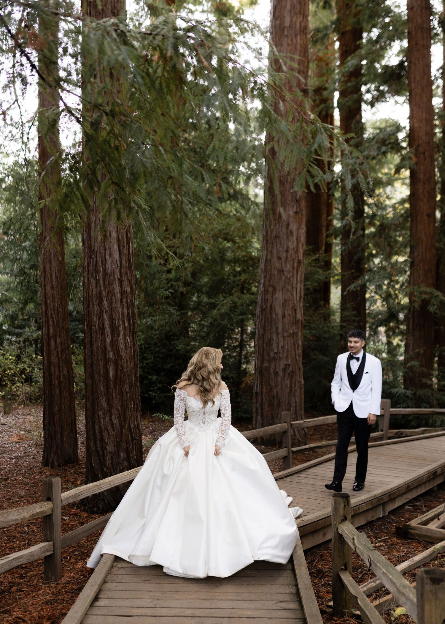 A bride in a ballgown and groom in white tuxedo pose on a wooden walkway in a redwood forest.