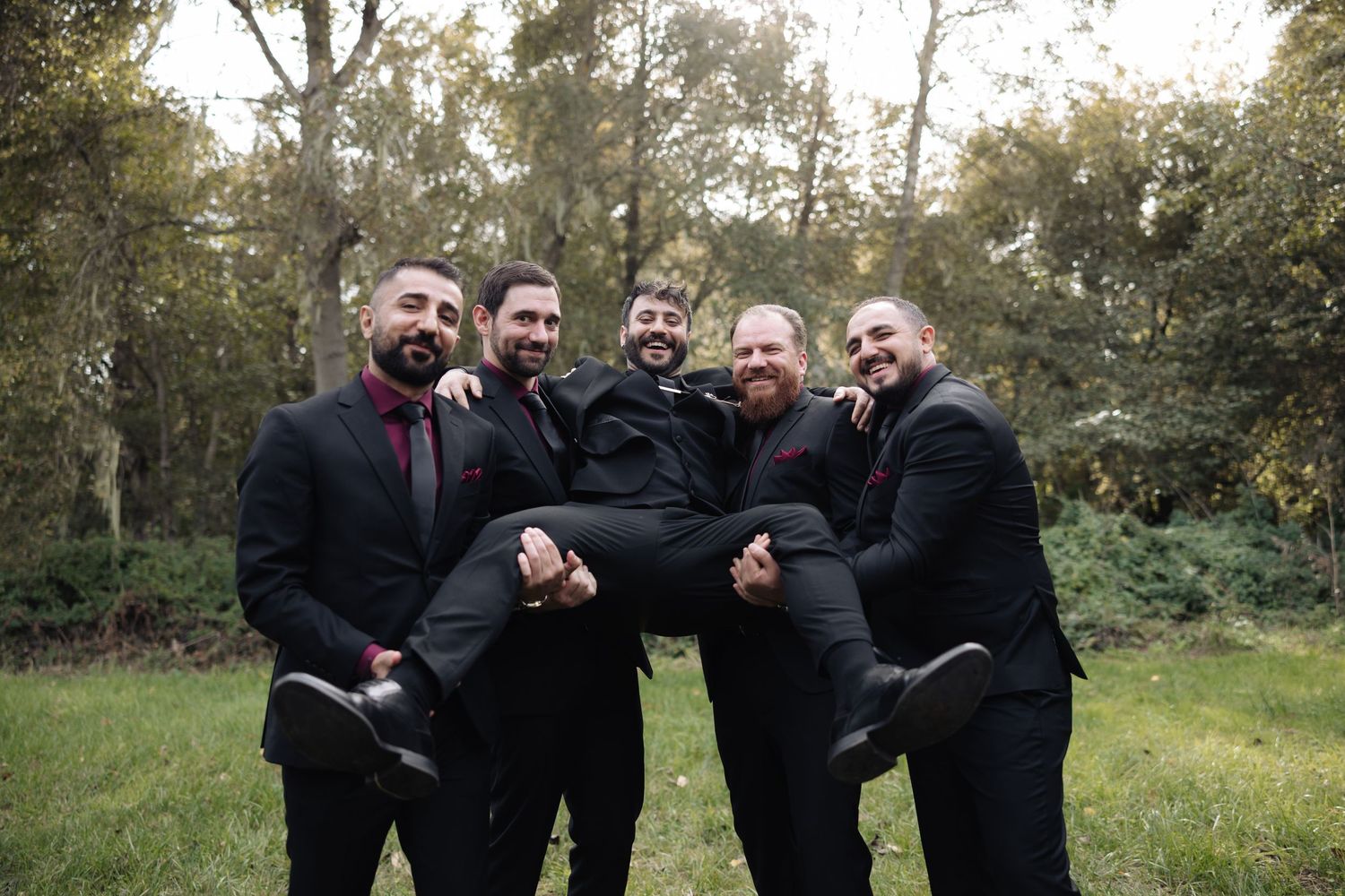 Groomsmen in black suits pose playfully for candid photos in a natural outdoor setting.