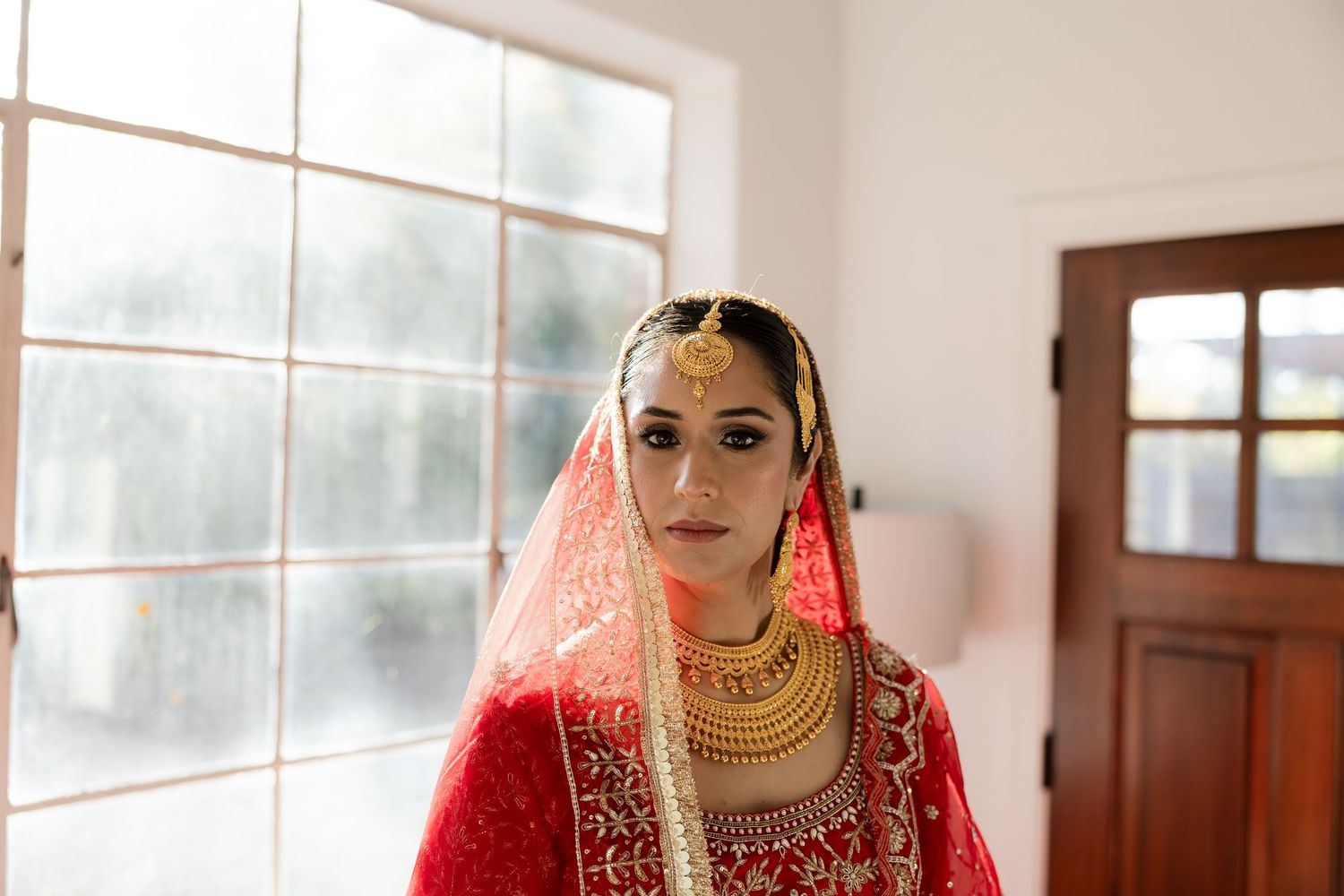 Traditional Indian bridal attire featuring red dress, gold necklaces and veil against window backdrop.