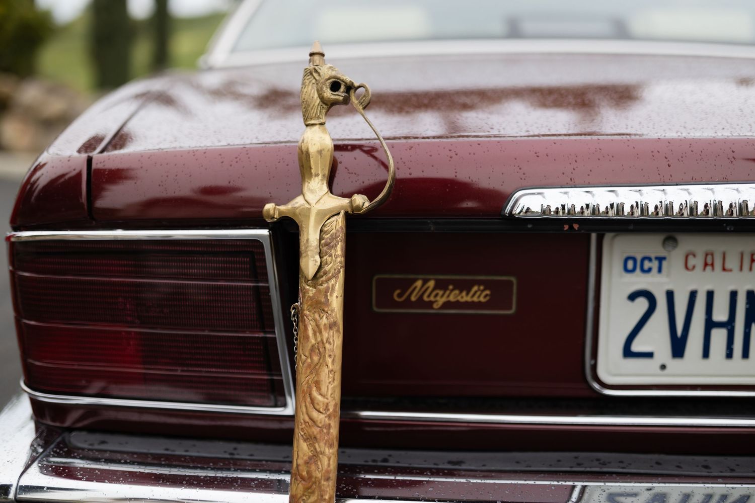 A decorative golden sword ornament mounted on the trunk of a vintage maroon Chevrolet Caprice Classic car.
