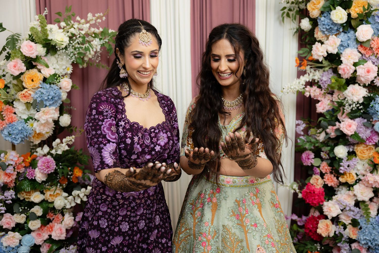 Women in elegant traditional outfits pose together against a floral backdrop at a festive celebration.
