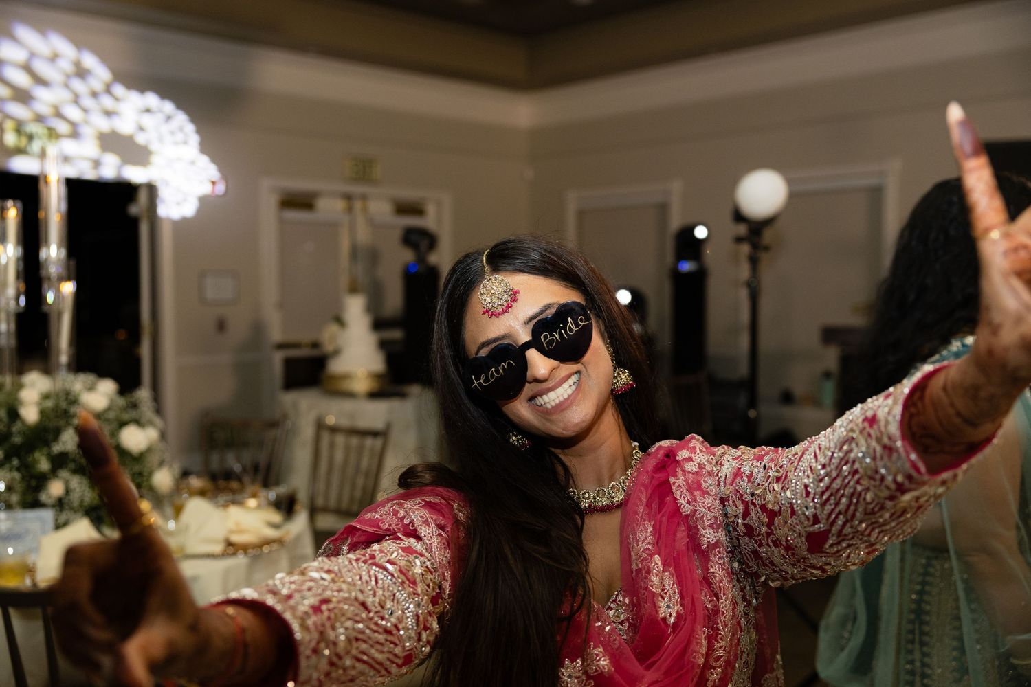 A group of people dancing and celebrating at an indoor wedding reception with decorative lighting and floral arrangements.