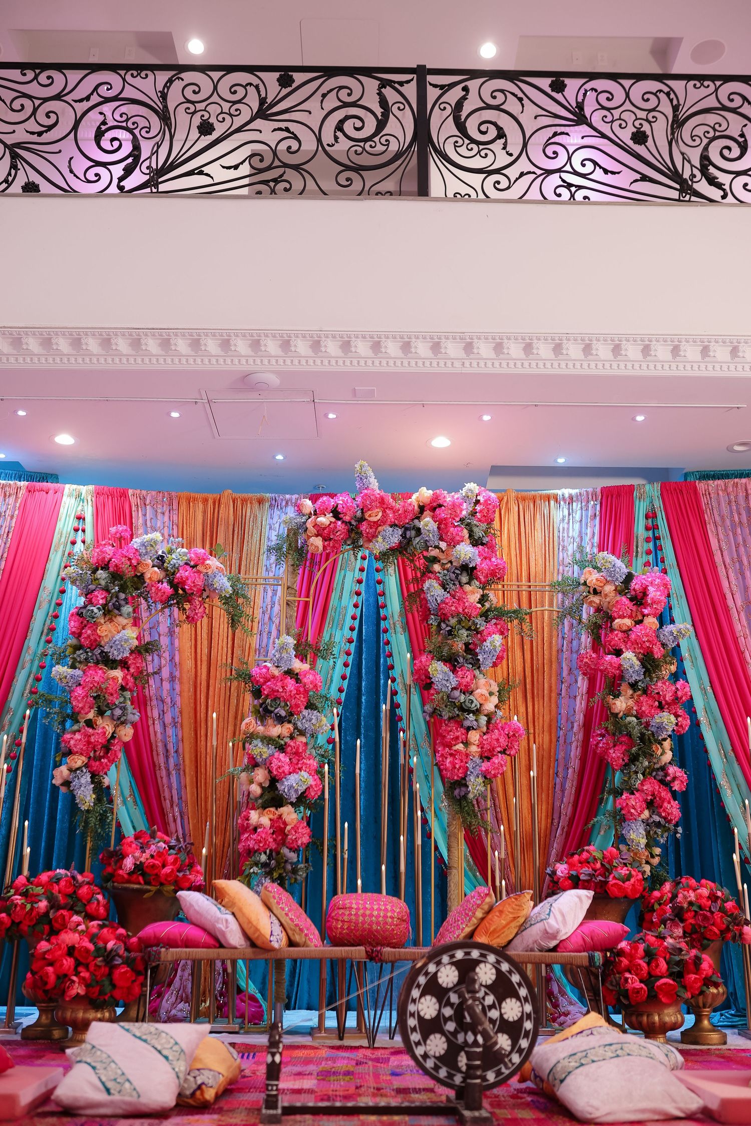 Colorful wedding stage decor with pink and turquoise drapes, floral arrangements, and plush cushions creates a vibrant ambiance.