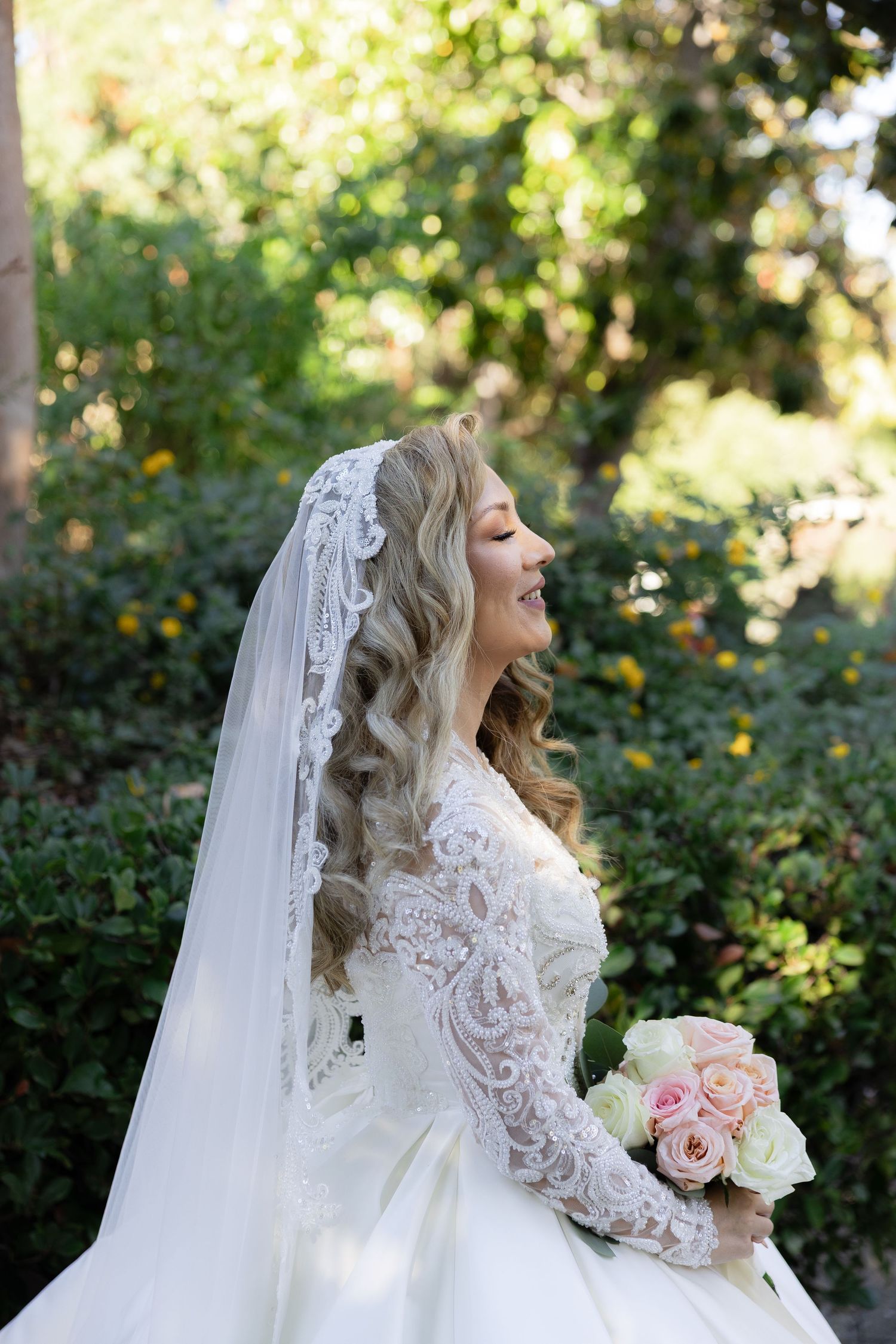 Elegant bridal portrait in a garden setting wearing a lace long-sleeve wedding gown with flowing veil.