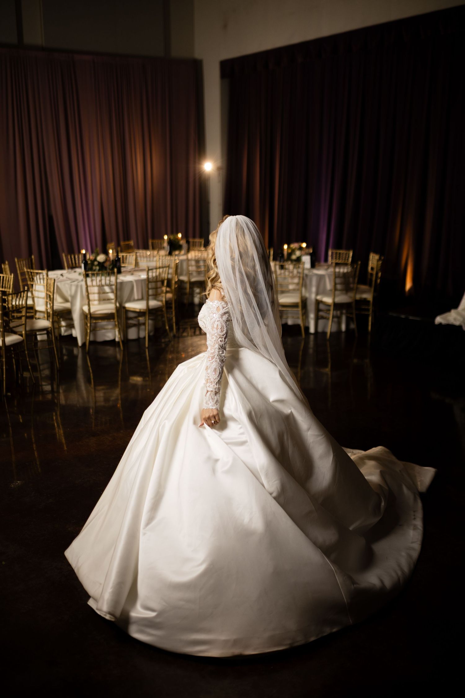 A side profile of a bride in a classic ball gown surrounded by gold chairs in a reception hall.