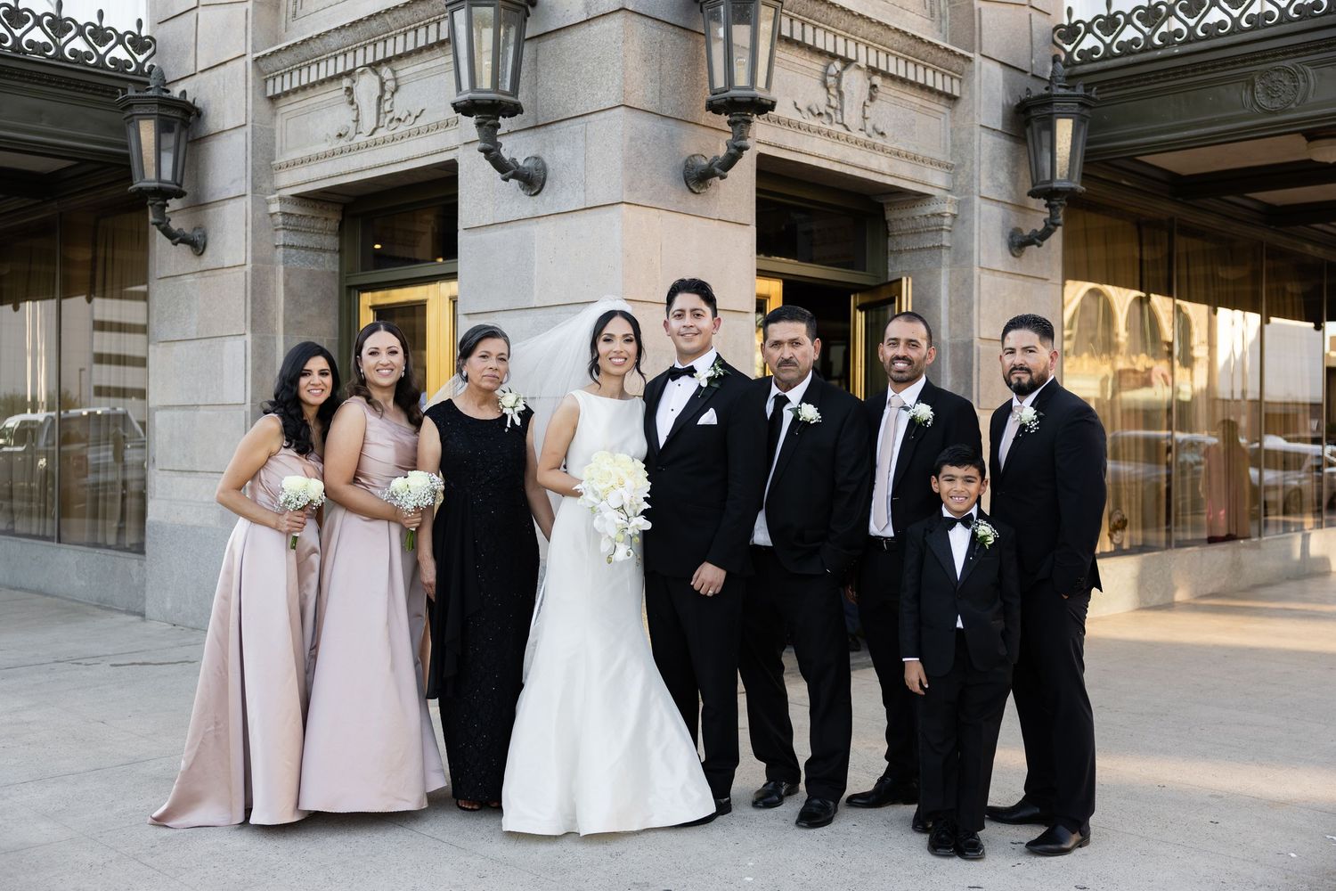 Wedding party in formal attire poses together outside historic building entrance.