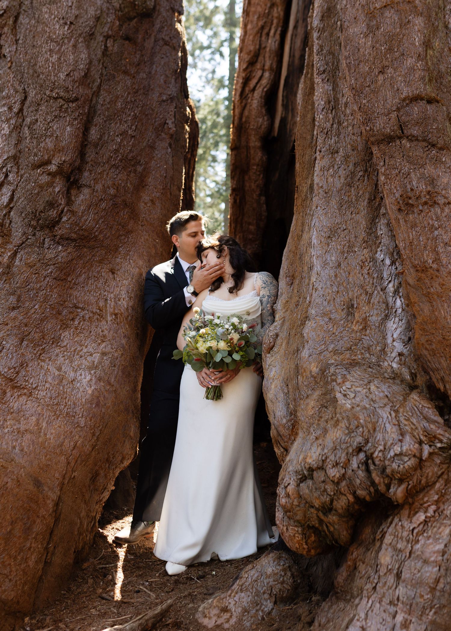 Bride and groom embrace between giant sequoia trees, bride holding wildflower bouquet, in a romantic forest wedding photo.