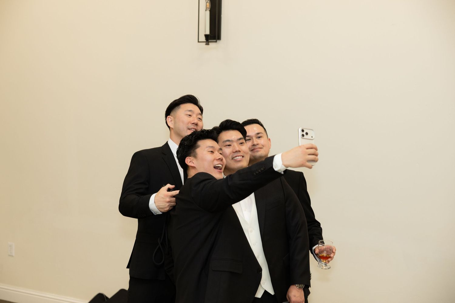 Four groomsmen in black suits take a fun group selfie together at a wedding celebration.