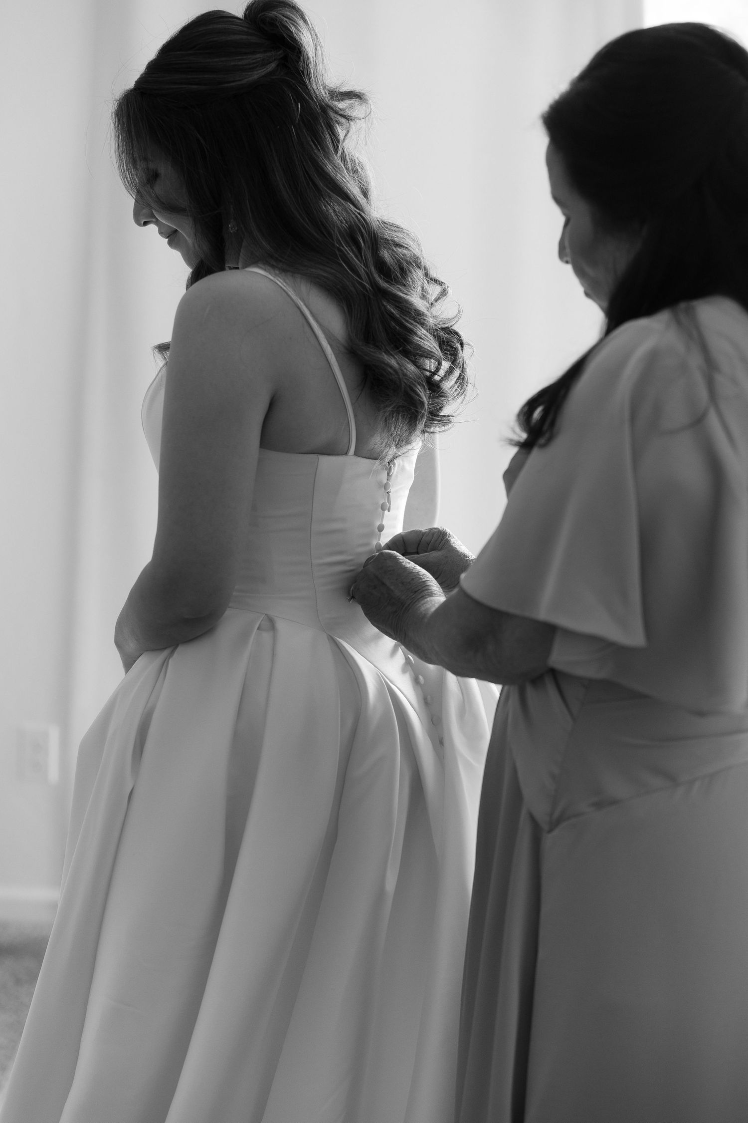 Black and white photo of a bride getting her wedding dress buttoned up by a helper before her ceremony.