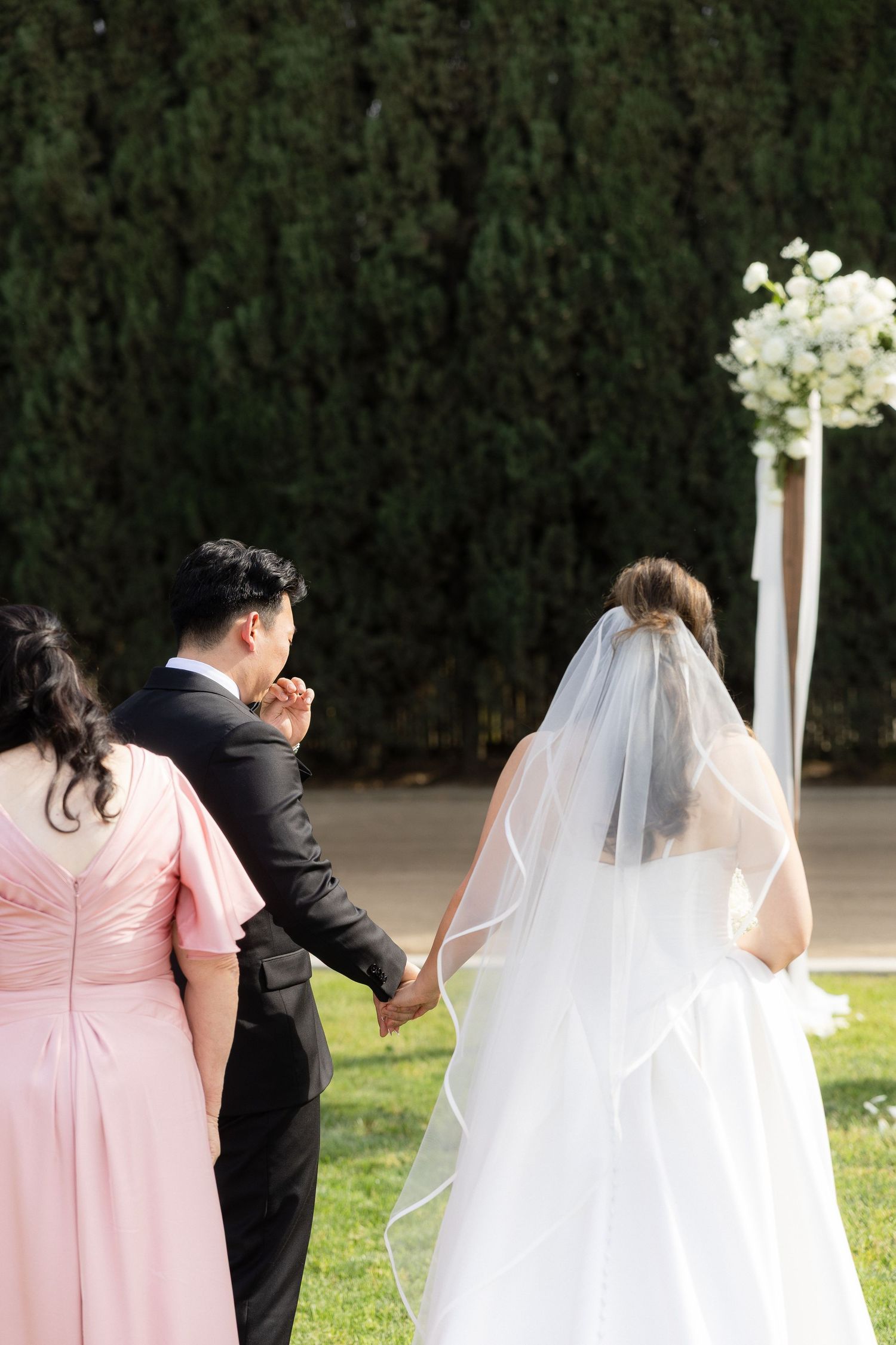 Bride in white veil walks with groom and woman in pink dress toward floral wedding arch.