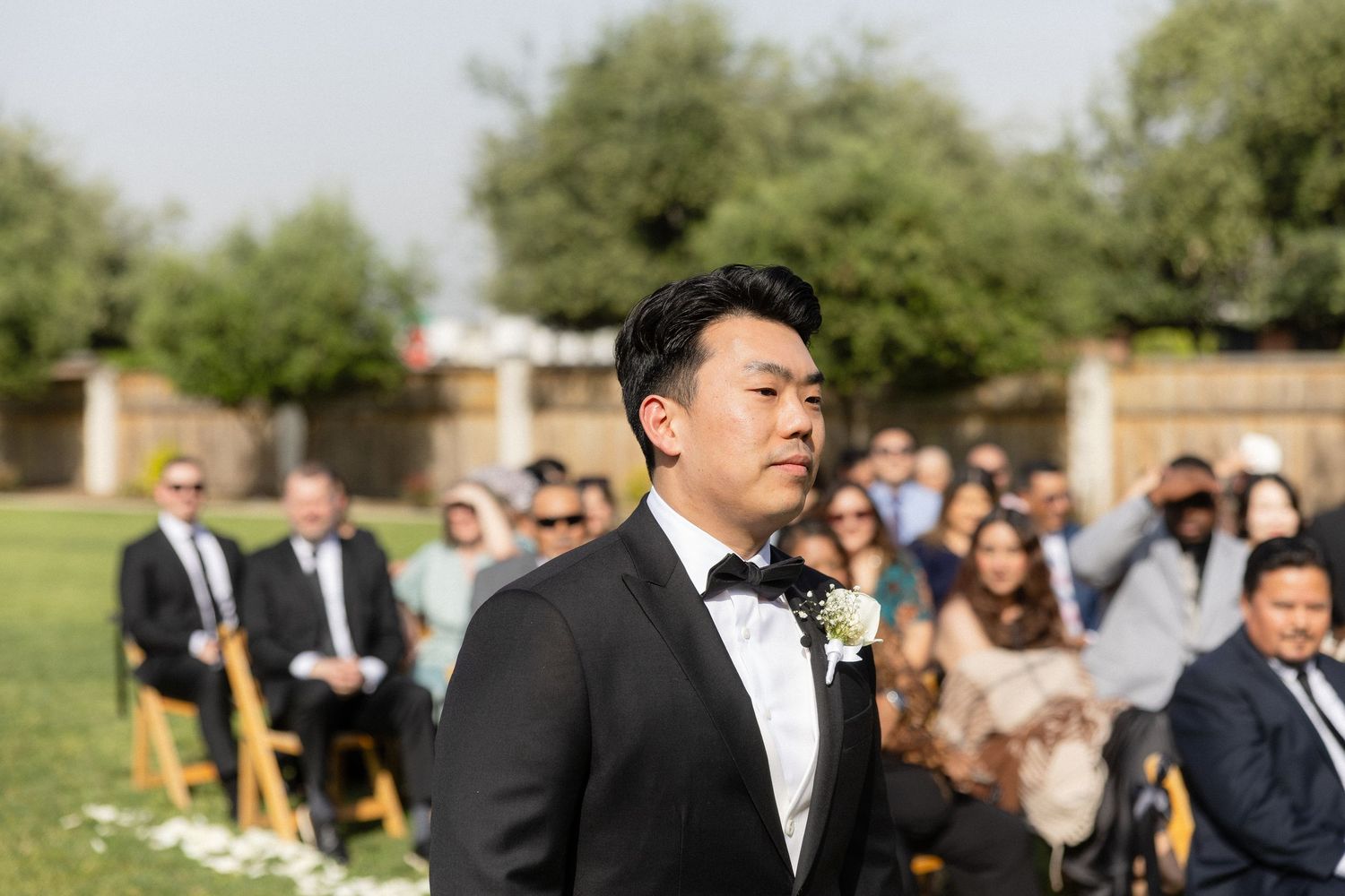 Groom in black tuxedo with boutonniere awaits bride at outdoor wedding ceremony with seated guests.