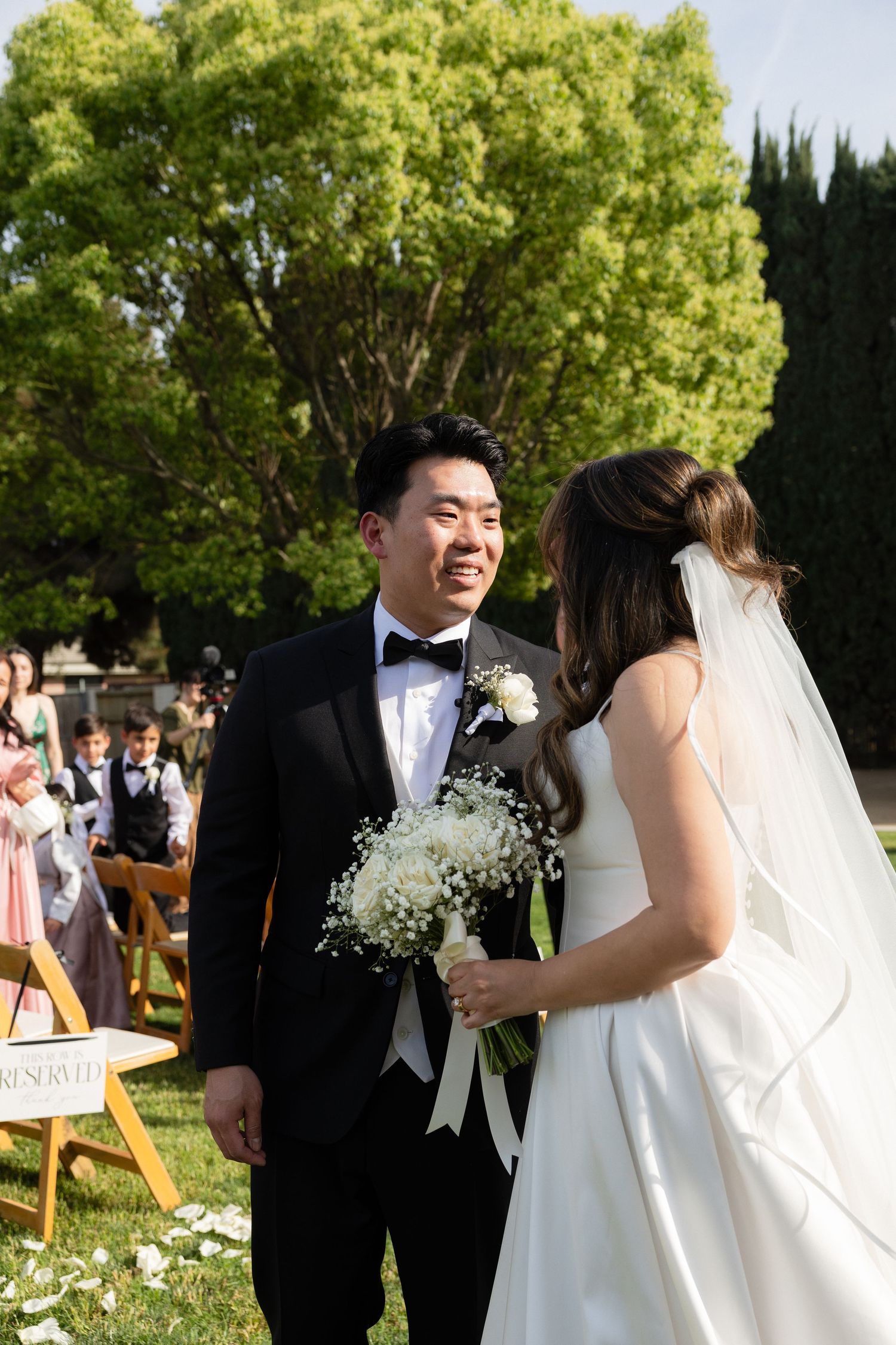 Groom in tuxedo smiles at bride in white gown holding bouquet during outdoor wedding ceremony.