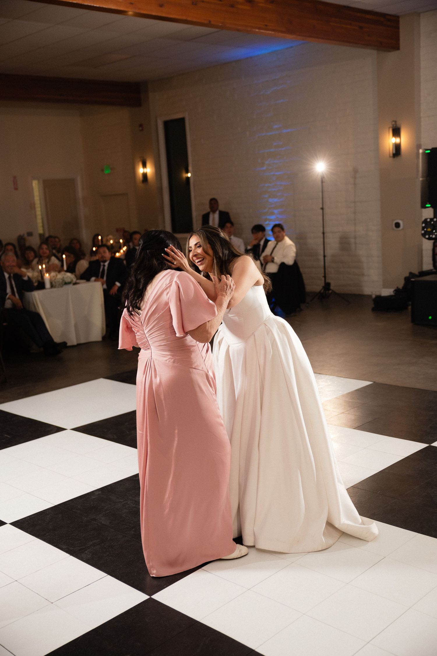 Bride in white gown shares emotional dance with bridesmaid in pink dress on checkered dance floor.