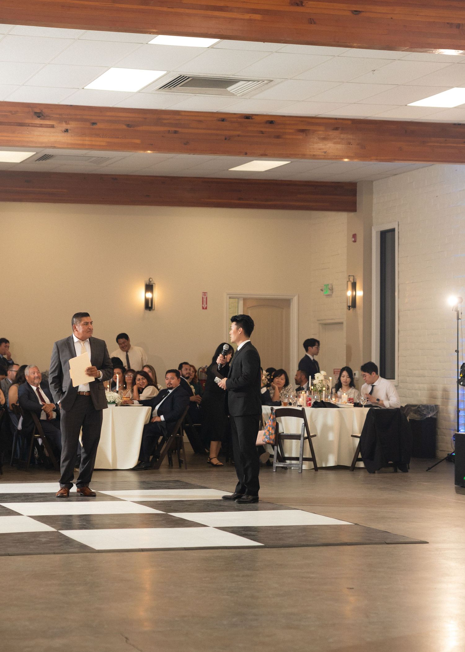 Groom and best man giving speeches on checkered dance floor at wedding reception.
