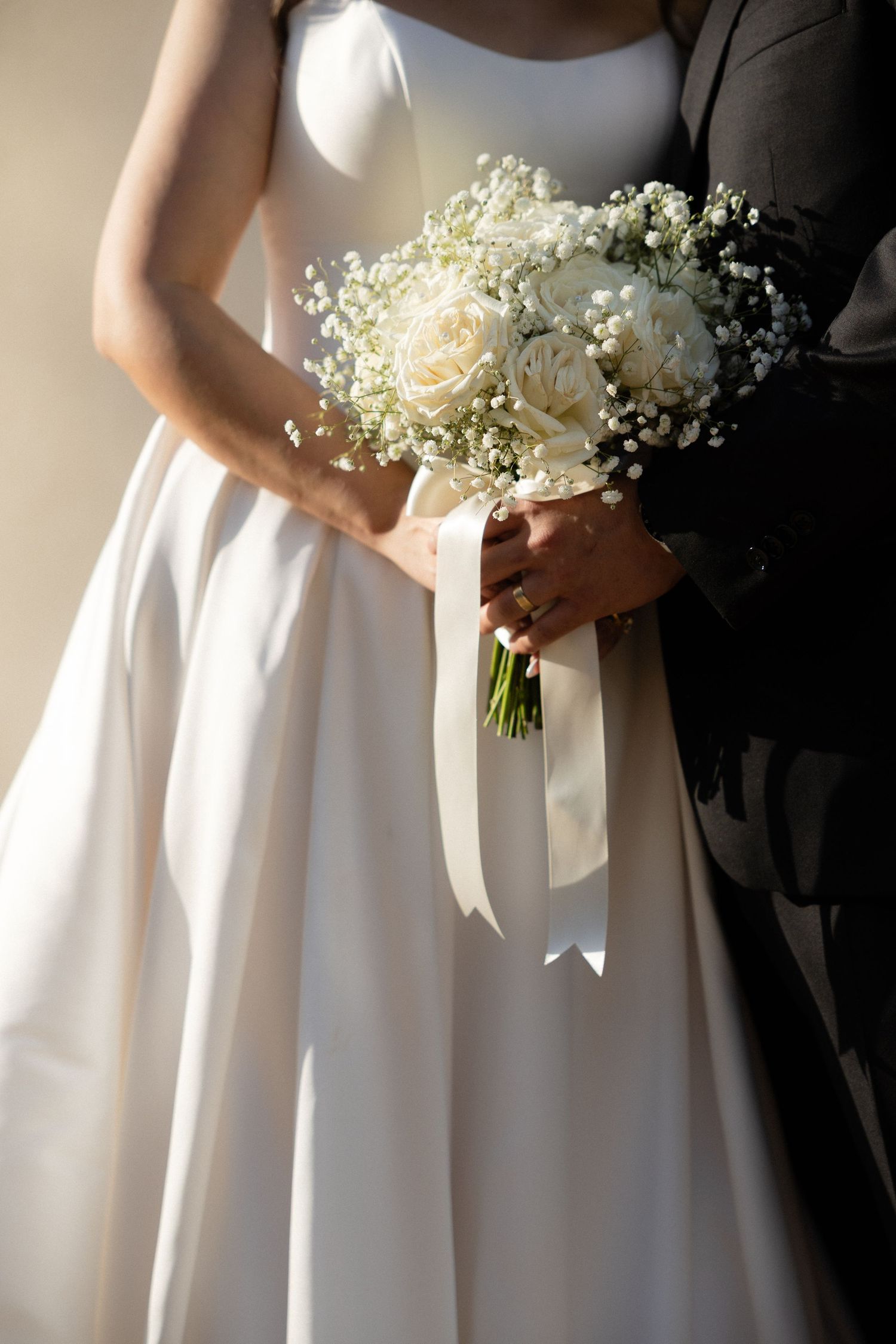 Bride in white gown holding elegant bouquet of white roses and baby's breath with satin ribbon beside groom in black suit.