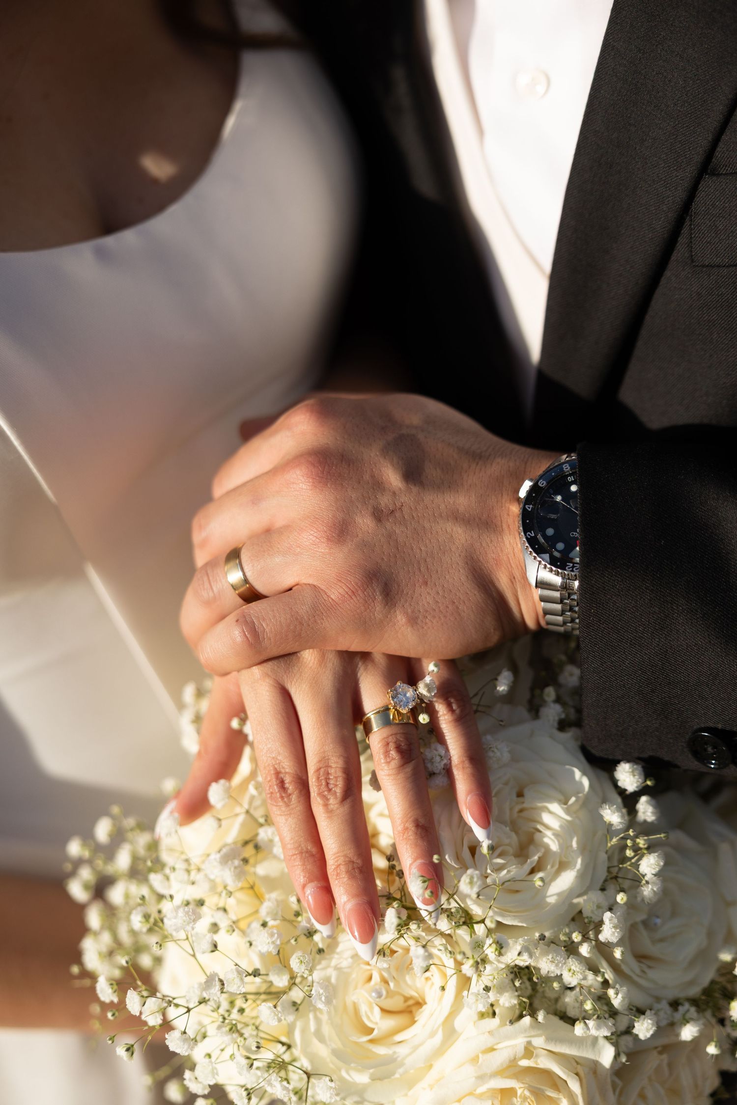 Newlywed couple displaying wedding rings and a luxury watch over a white rose bridal bouquet.