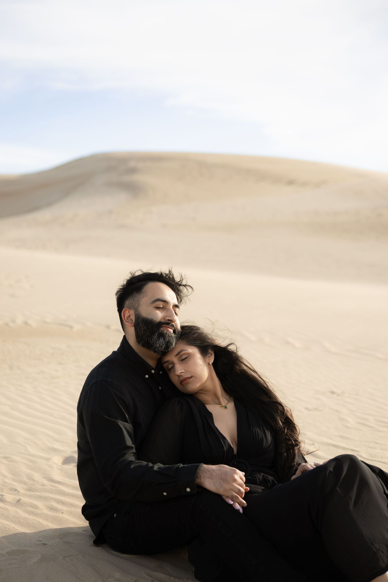A couple dressed in black sits intimately together on desert sand dunes under a soft cloudy sky.