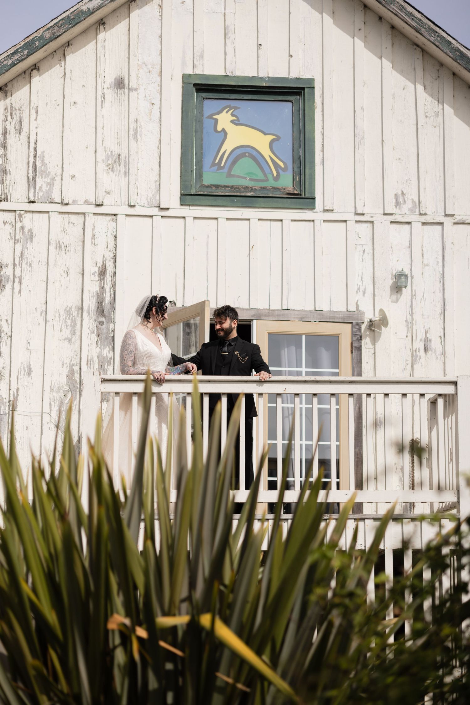 A couple stands on a rustic white barn balcony beneath a decorative yellow deer sign on a sunny day.