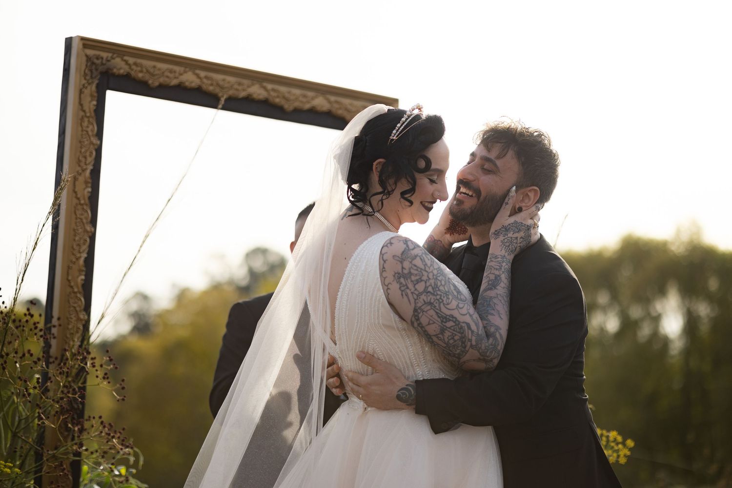 Couple embraces and laughs together under rustic wooden arch during golden hour wedding ceremony.