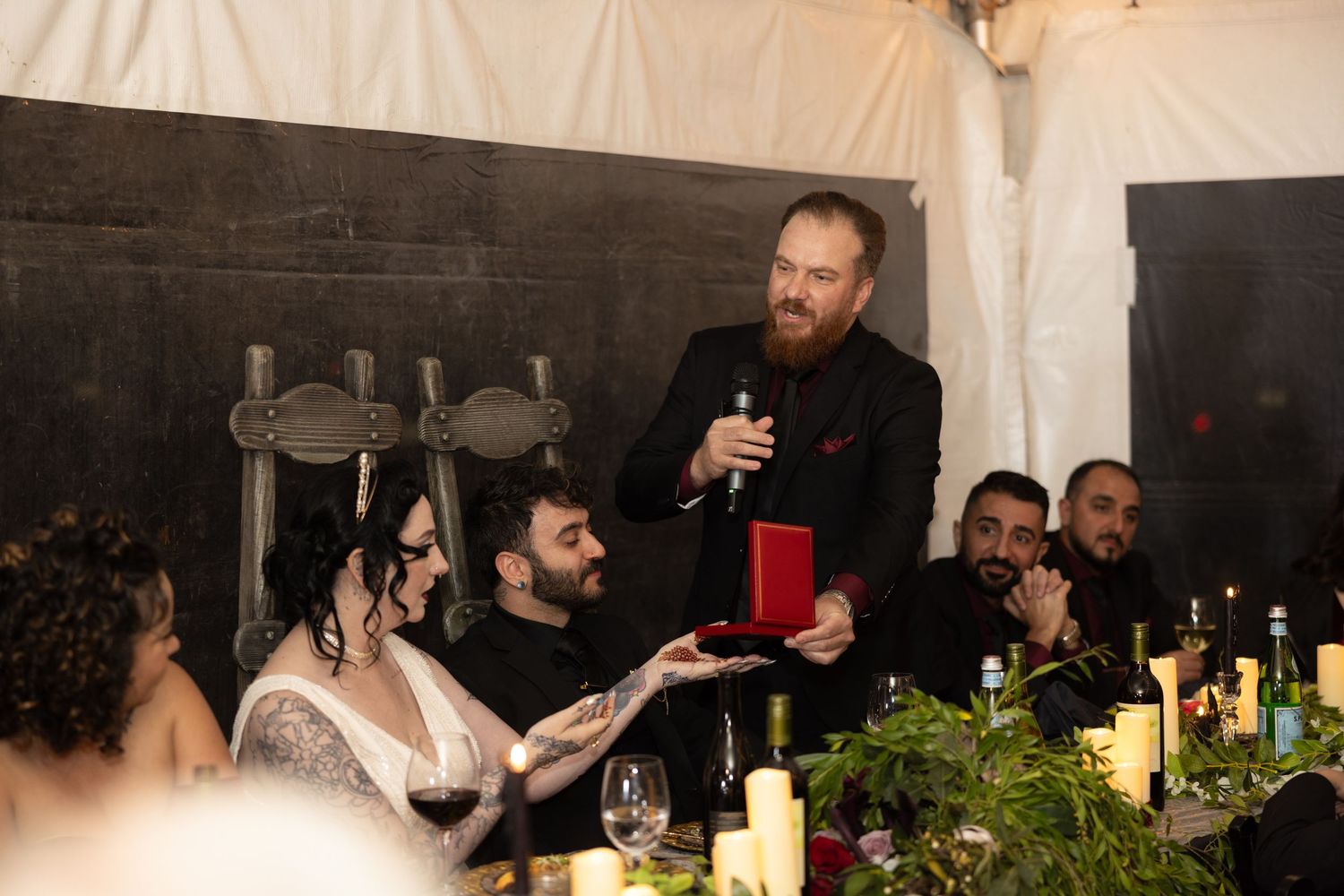 Wedding reception guests celebrate with drinks and dancing under warm string lights in a rustic venue.