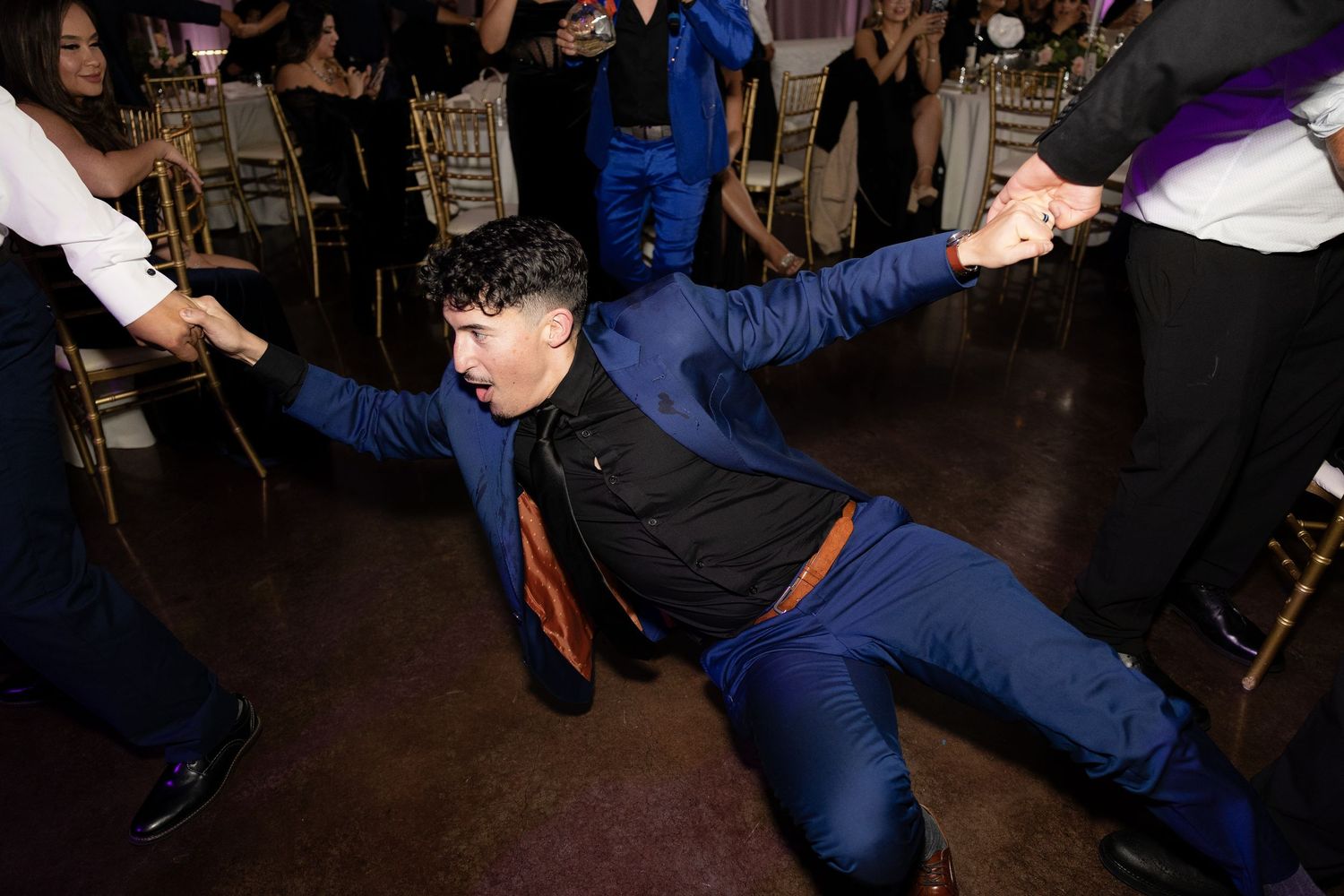 A wedding guest in a blue suit strikes a dramatic dance pose on the reception floor.