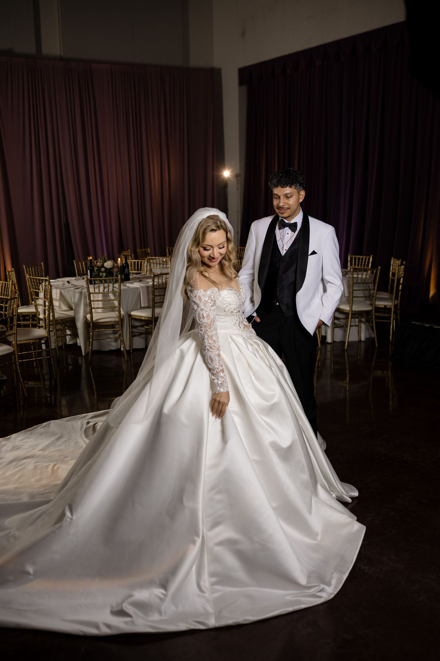 A bride in a luxurious white ballgown poses with her groom in an elegantly lit reception space.