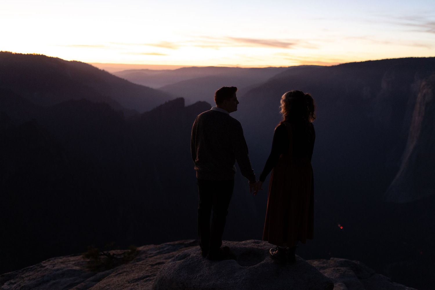 Silhouetted couple holding hands while standing on mountain overlook at dusk.