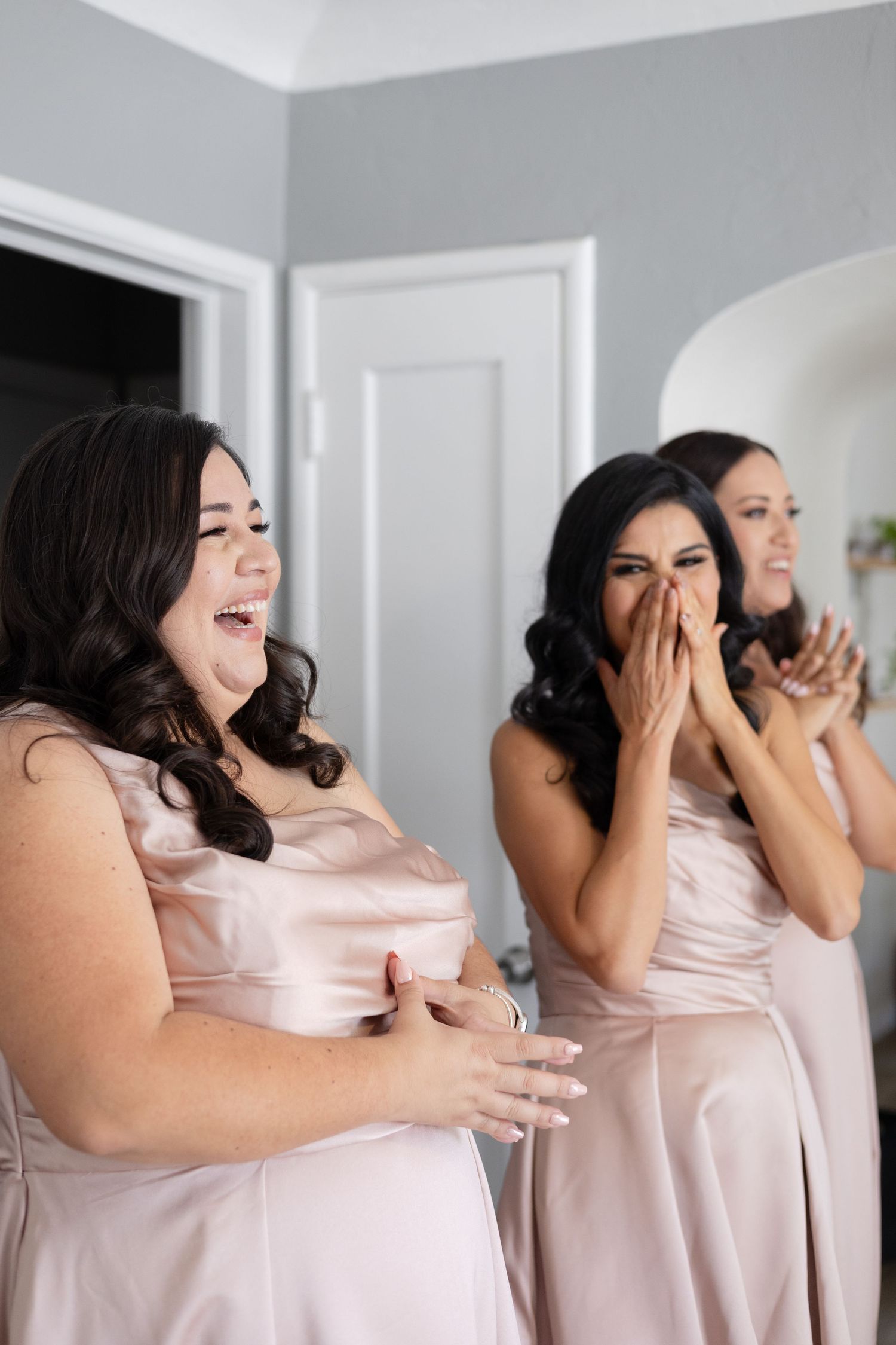 Bridesmaids in pink dresses react with laughter and excitement during a candid wedding moment.