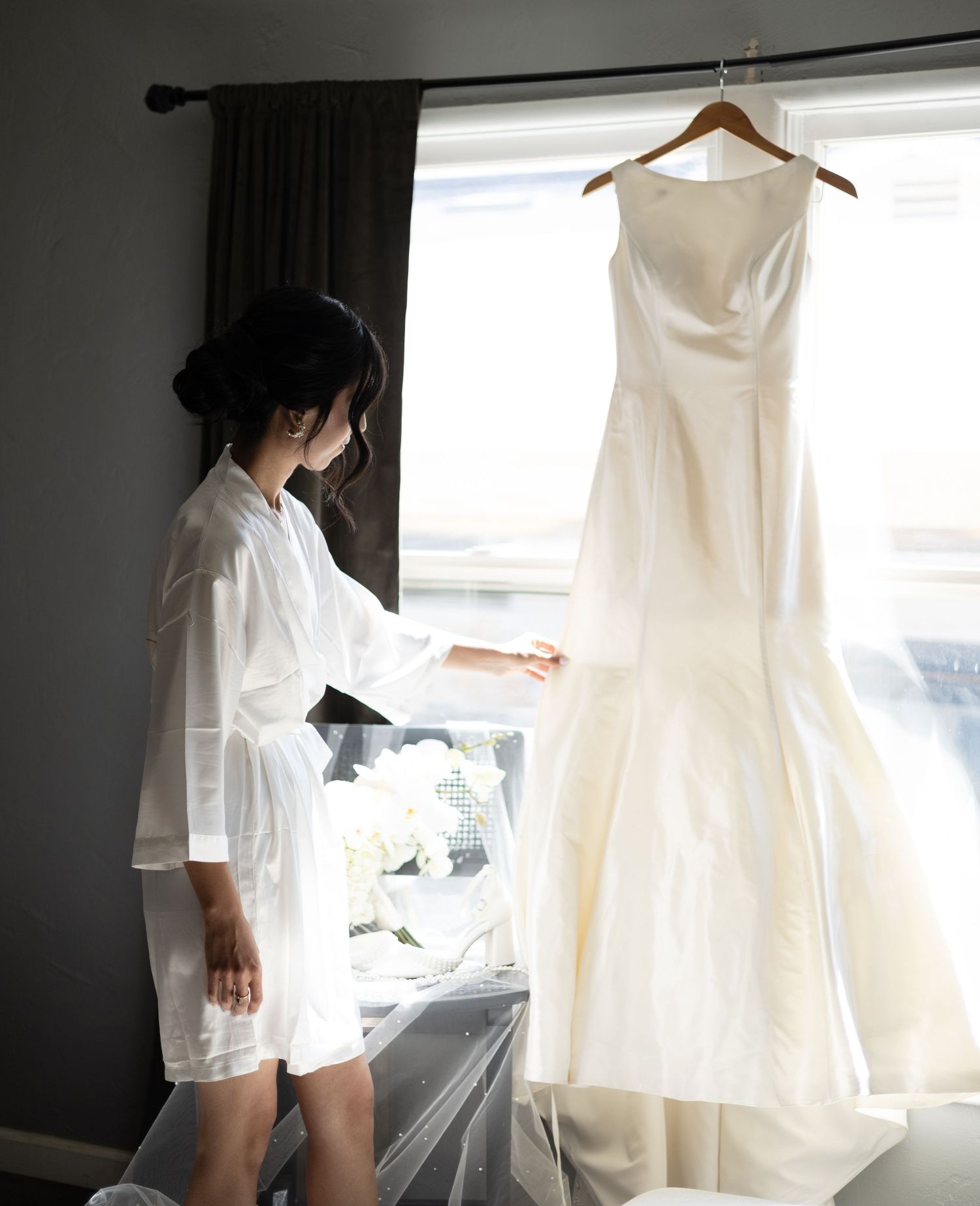 Wedding dress hanging by a sunlit window while someone in white robe stands nearby.