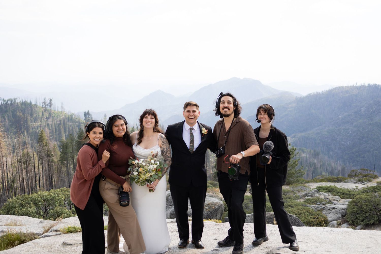 Wedding couple poses with guests on rocky mountain overlook with forested valley and misty peaks backdrop.
