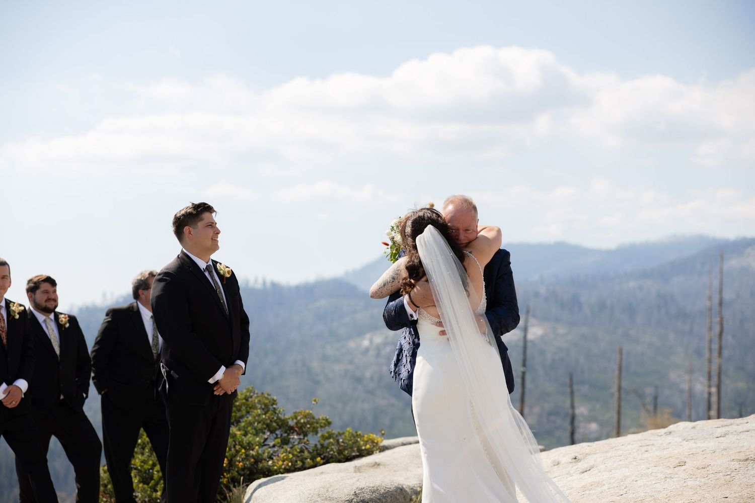Bride and groom share first kiss at outdoor mountain wedding ceremony with groomsmen watching scenic forest backdrop.