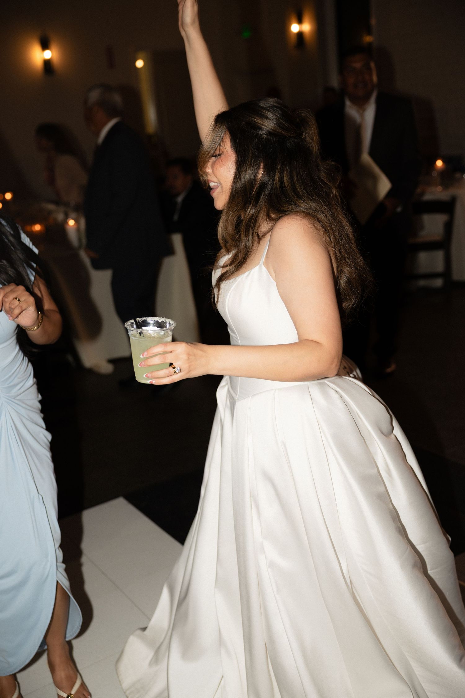 Bride in white satin gown dancing with raised arm holding a cocktail at reception.