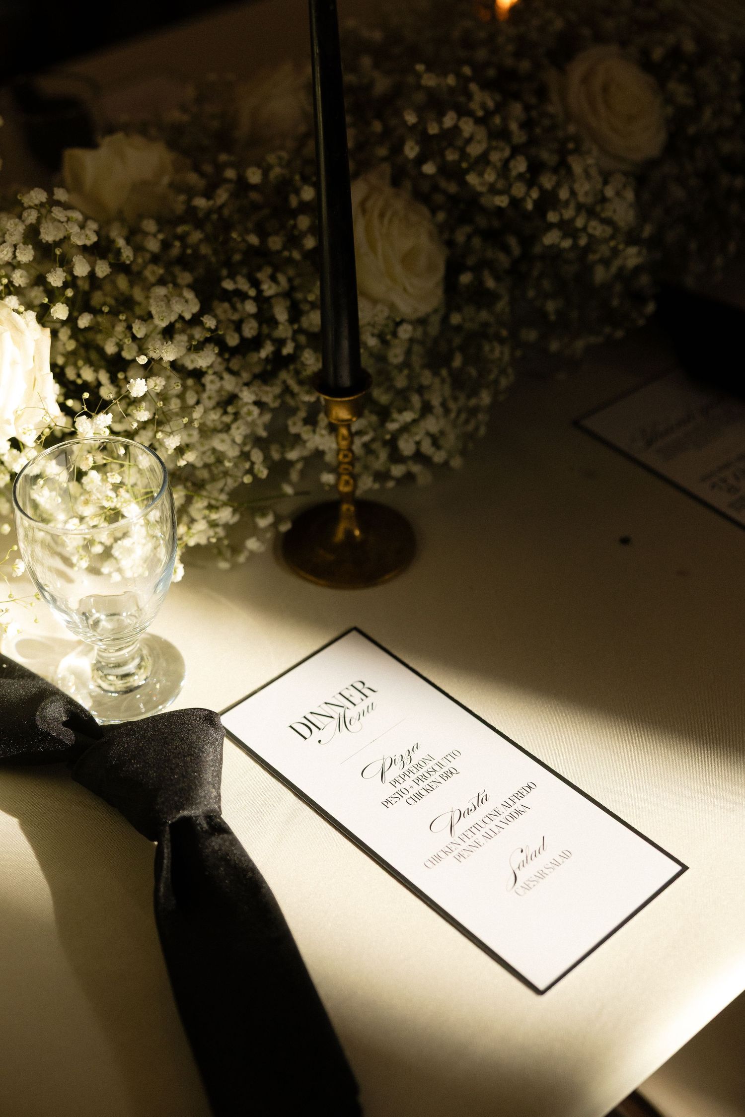 Elegant wedding reception dinner menu card with black ribbon, crystal glass, white roses, and baby's breath centerpiece on white tablecloth.