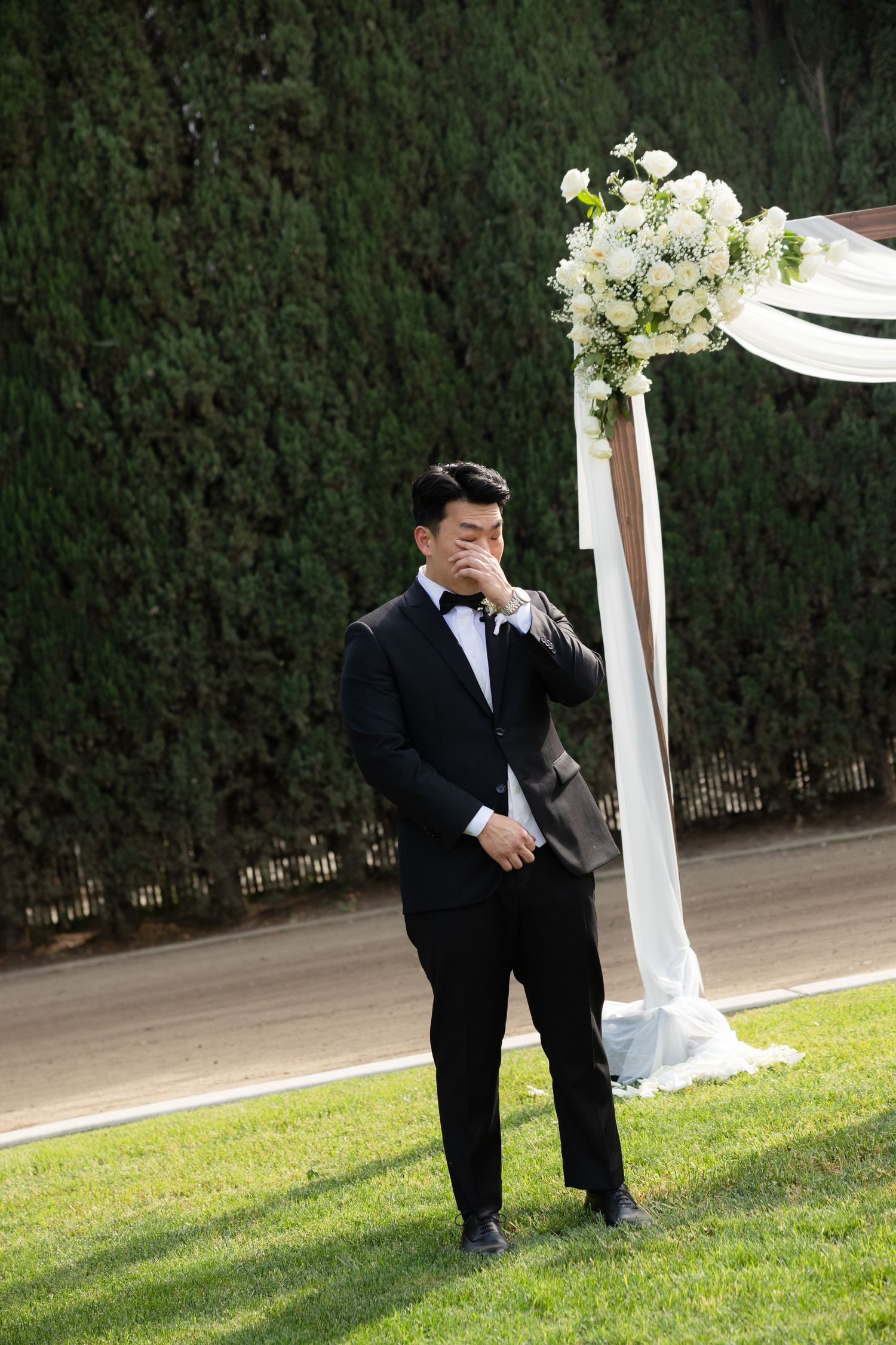 Emotional groom in black tuxedo wiping tears beside floral wedding arch on lawn.