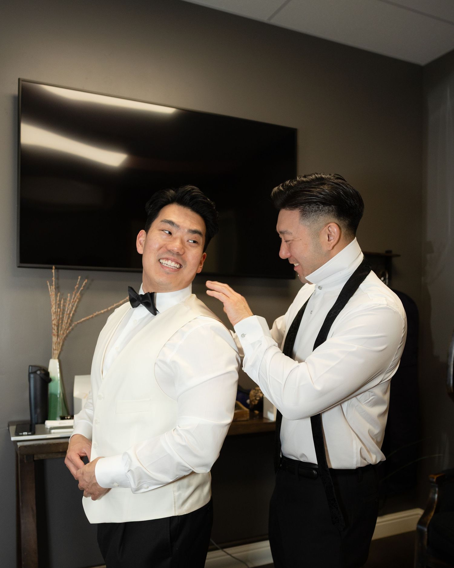 A groomsman adjusts the bow tie of a smiling groom in a white tuxedo jacket before a wedding ceremony.