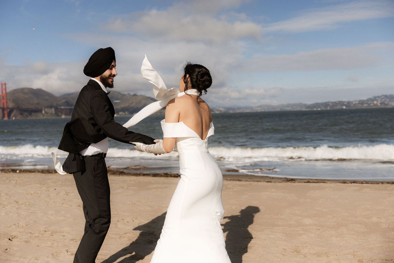 A couple in wedding attire runs and dances playfully along a scenic beach with waves and mountains in the background.