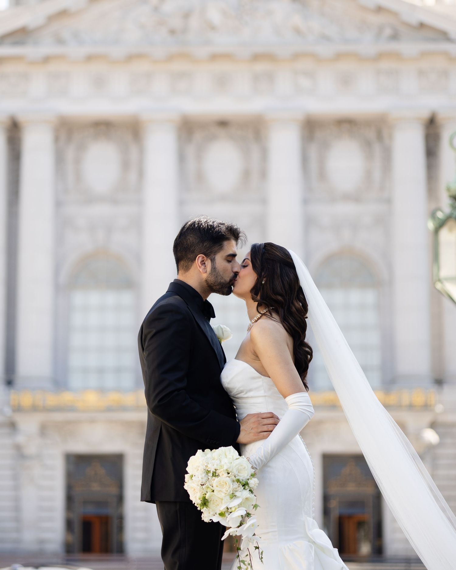 A romantic wedding couple shares an intimate kiss outdoors against a grand architectural backdrop.