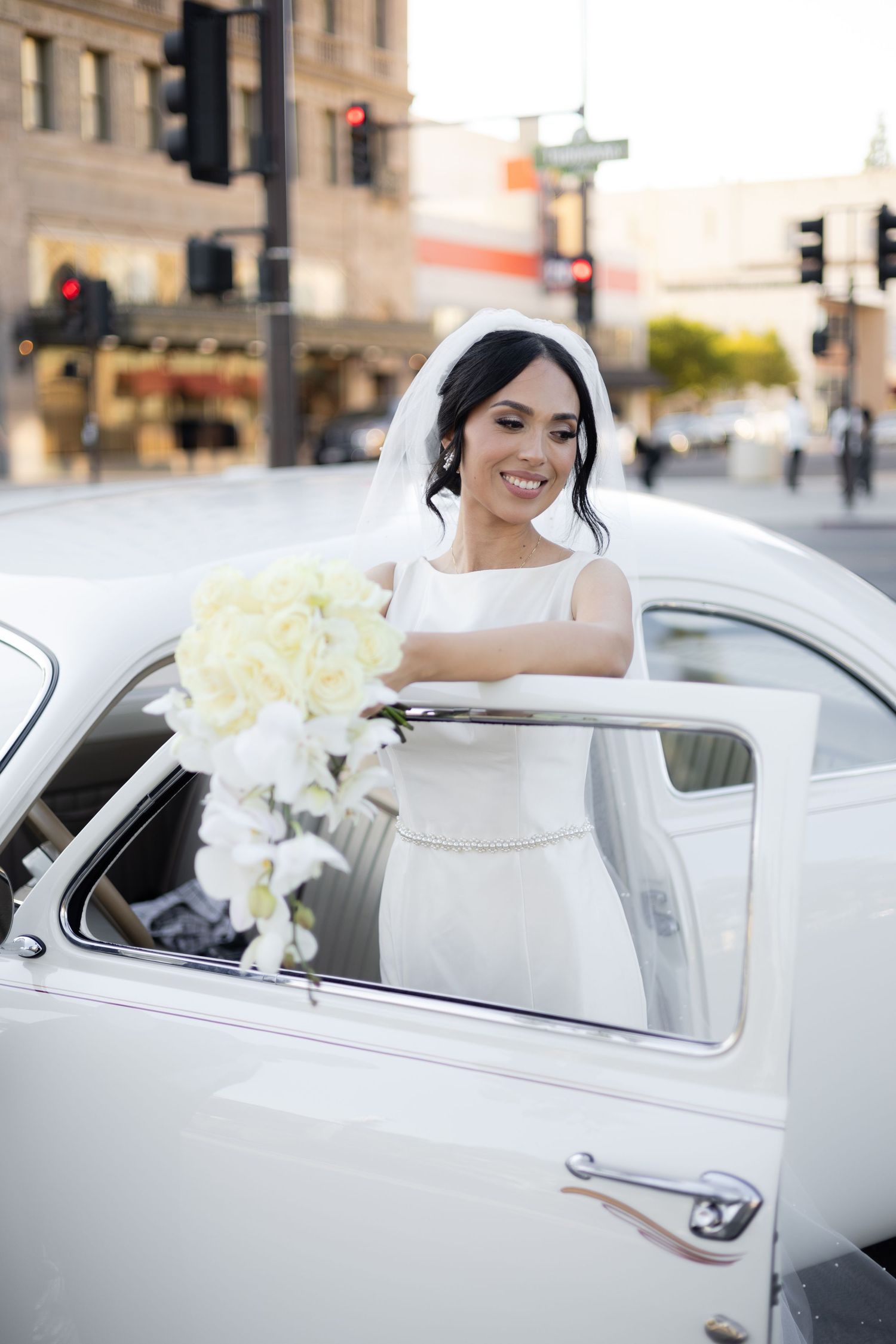 Wedding couple poses with classic white car decorated with elegant white floral arrangements.