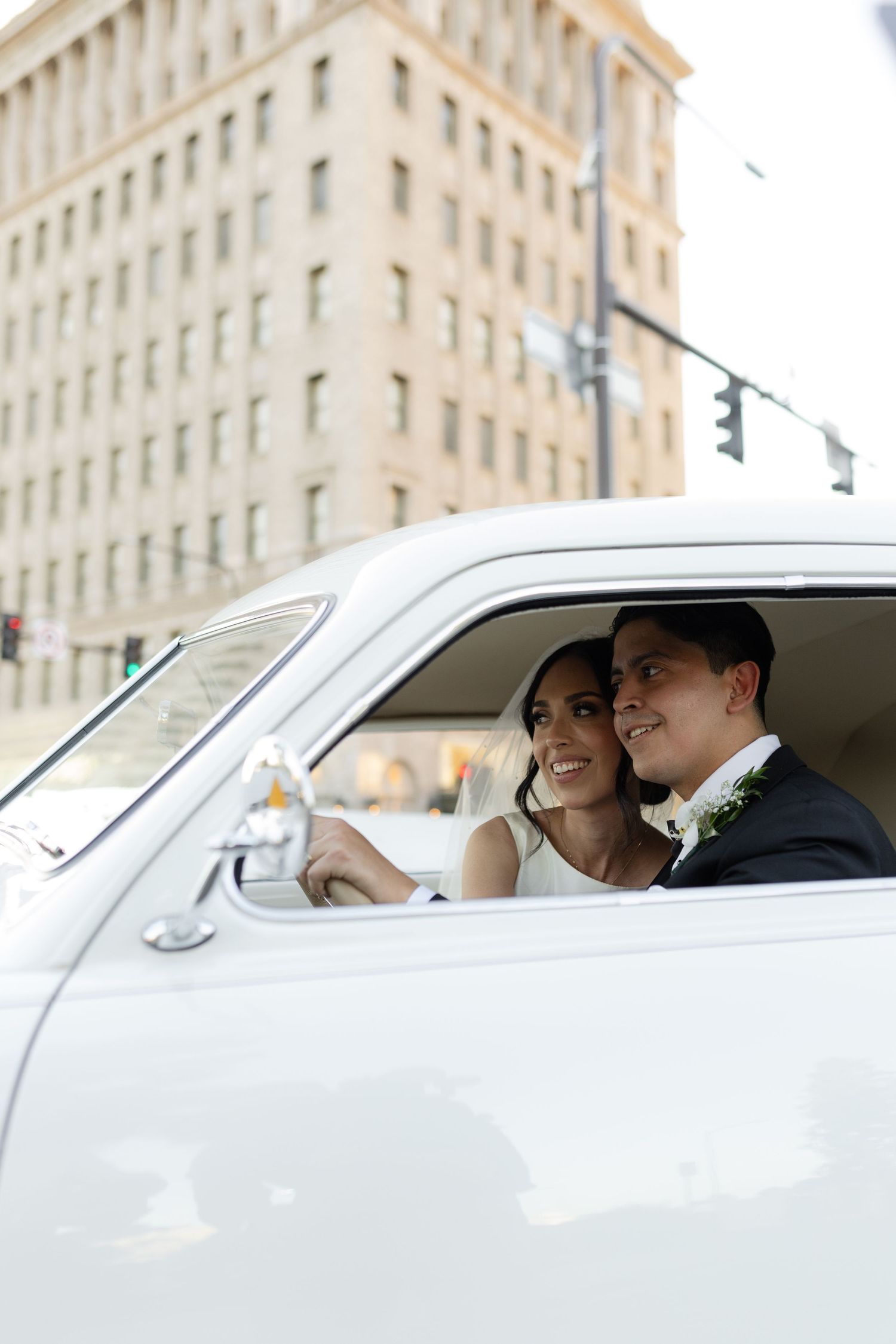 A couple shares a tender moment in a classic white car with an elegant architectural backdrop.
