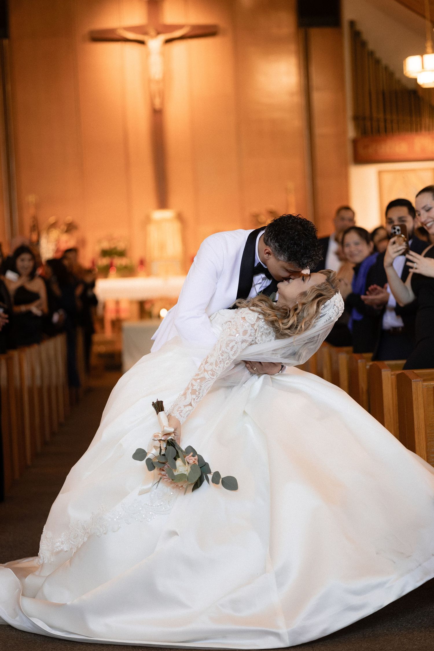 Dramatic wedding kiss in church aisle with guests watching and crucifix glowing behind.