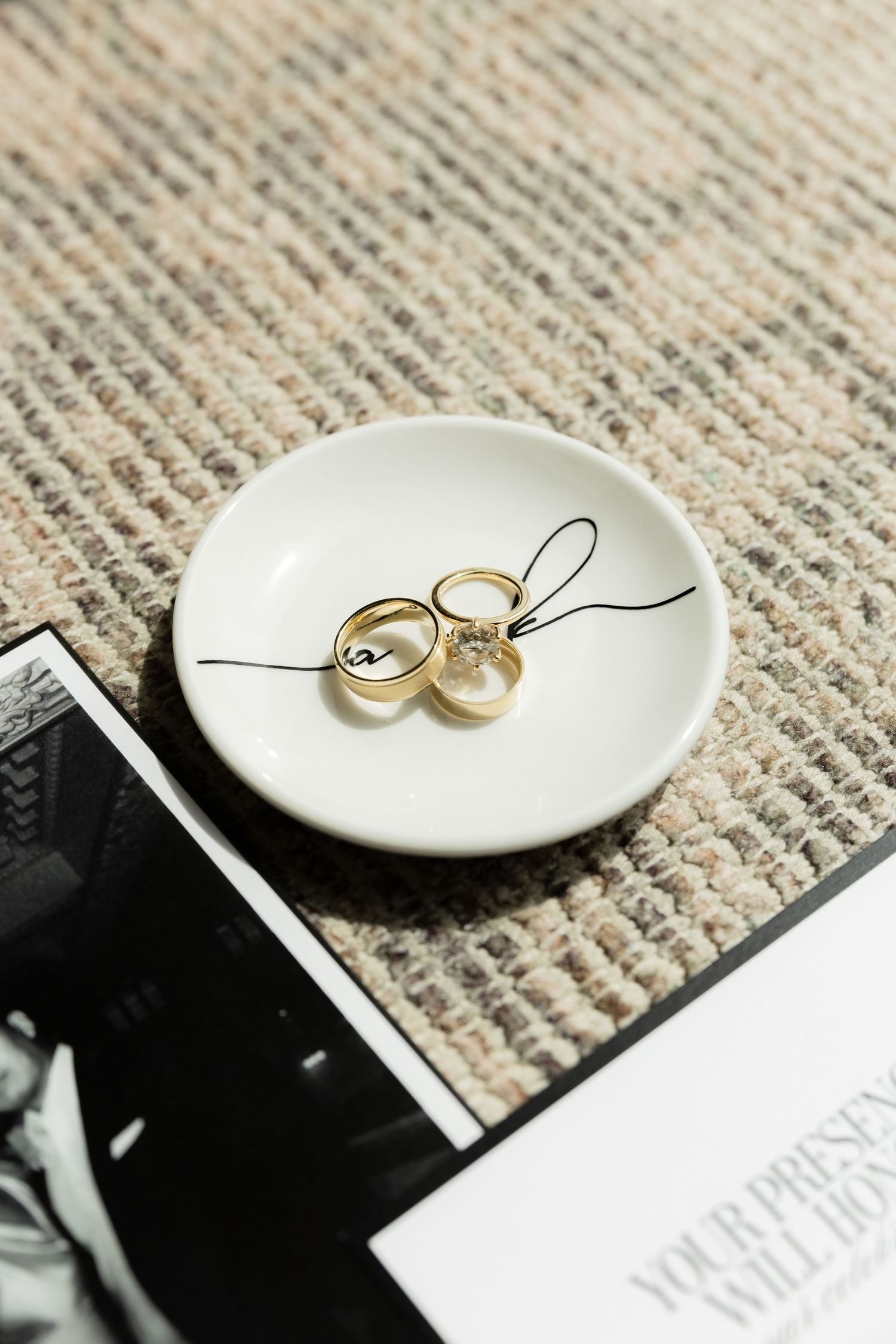 Gold wedding rings on a white ceramic ring dish atop a woven rug beside black and white photos.